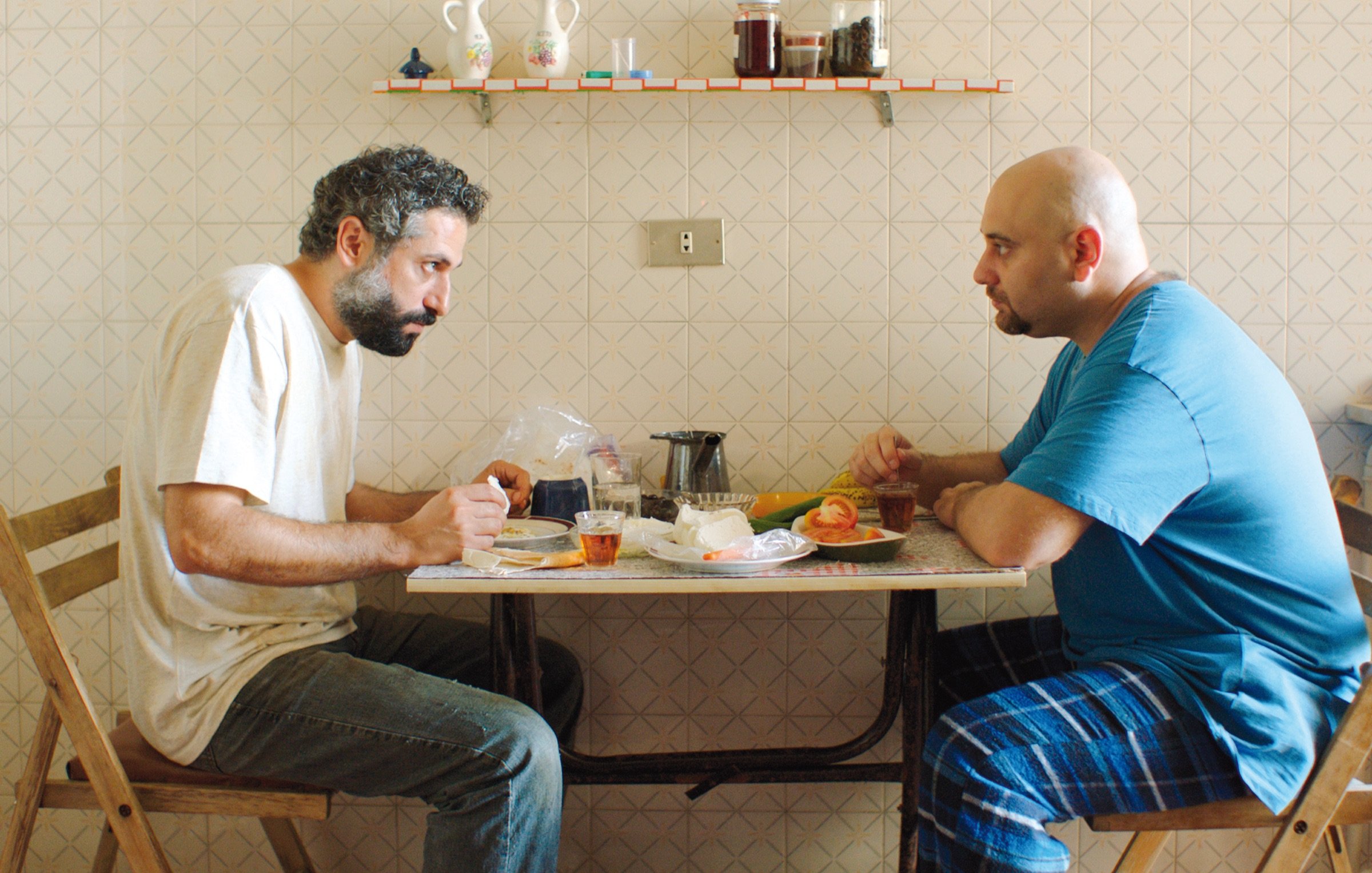 L'image montre deux hommes assis à une table dans une pièce au décor simple. Ils semblent en pleine conversation, chacun portant une expression sérieuse. Sur la table, il y a des assiettes, des verres et des aliments. L'un des hommes porte un t-shirt gris, tandis que l'autre est vêtu d'un t-shirt bleu. La pièce est éclairée de façon naturelle, et le mur est décoré d'un motif en damier. L'ambiance semble à la fois intime et chargée.