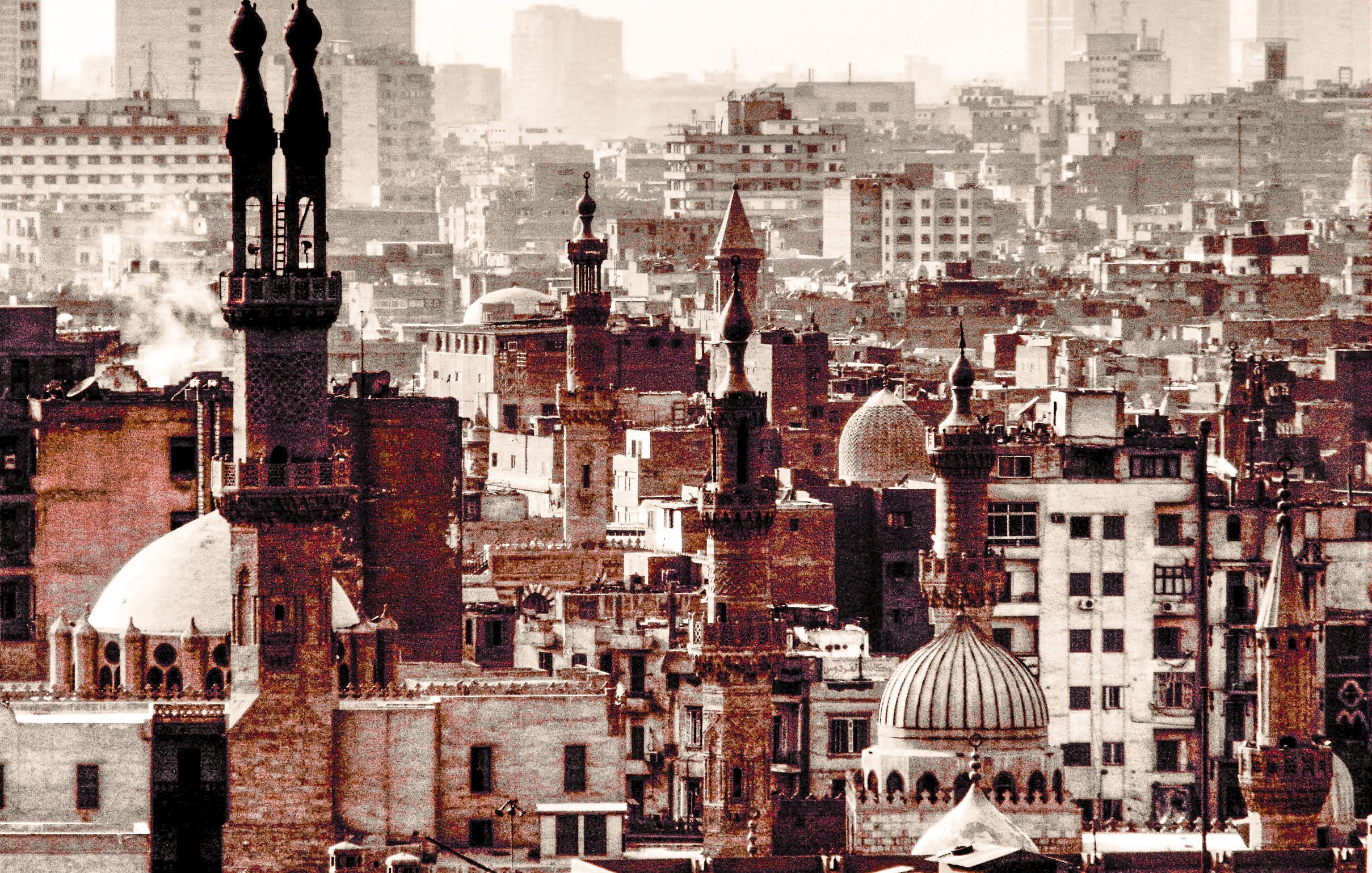 L'image montre un paysage urbain d'une grande ville, probablement au Moyen-Orient. On y distingue plusieurs minarets et dômes de mosquées, reflétant une architecture historique et religieuse. Les bâtiments sont de styles variés, certains ayant des tours élancées. La scène est baignée dans des teintes sèches, ce qui donne une ambiance nostalgique. En arrière-plan, on aperçoit des gratte-ciel modernes, créant un contraste avec l'architecture ancienne au premier plan. Le brouillard donne une impression de profondeur à l'image, soulignant l'intensité de la vie urbaine.