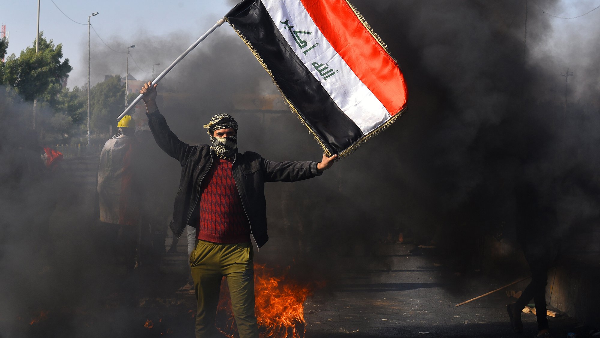L'image montre une scène de manifestation en Irak. Un homme, portant un couvre-visage et des vêtements sombres, brandit un drapeau irakien. En arrière-plan, on peut voir des nuages de fumée noire, suggérant des feux ou des barricades sur la route. L'atmosphère semble tendue, indiquant une situation de protestation.