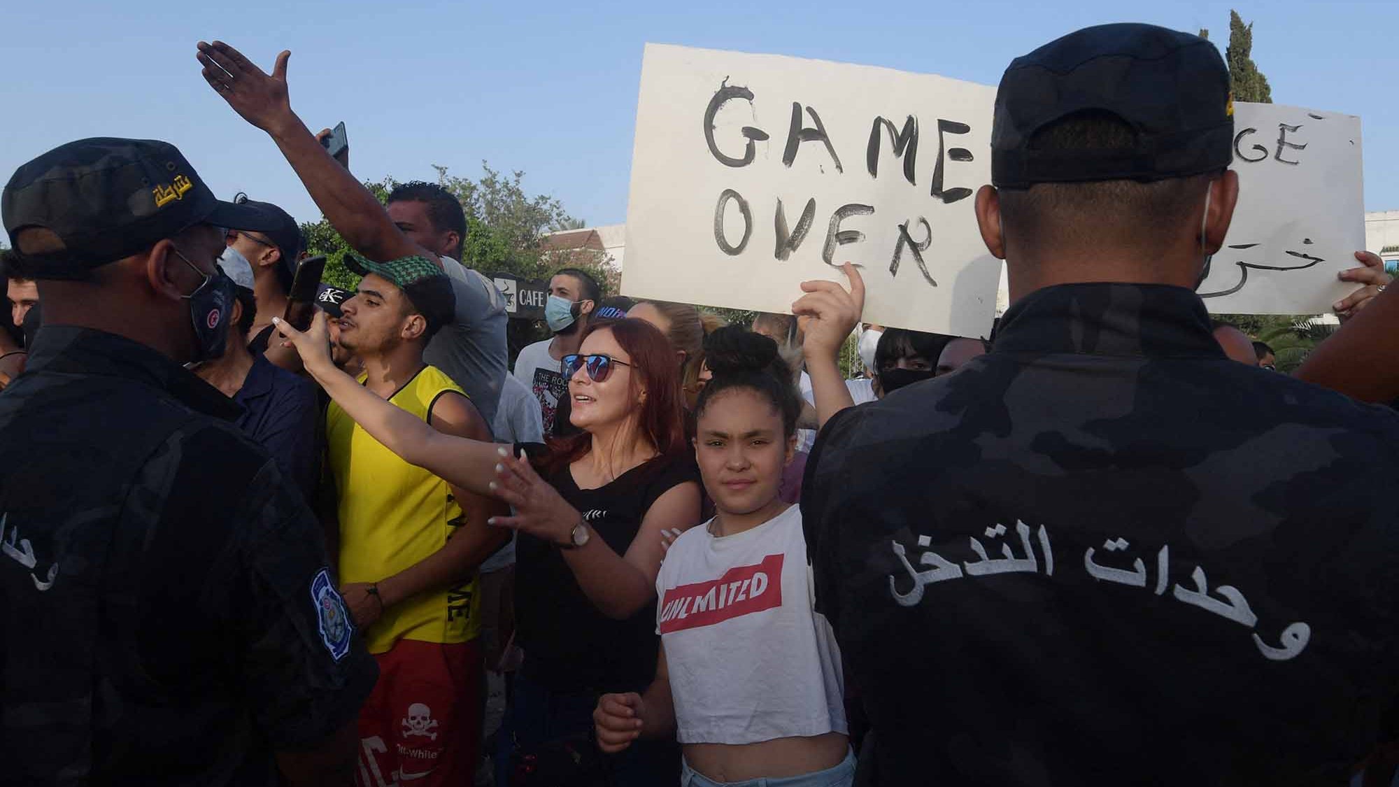 Dans l'image, on peut voir une scène de manifestation avec un groupe de personnes se rassemblant. Certains manifestants tiennent des pancartes, dont l'une indique "GAME OVER". Des policiers en uniforme, faisant face aux manifestants, semblent surveiller la situation. L'ambiance est tendue, avec des expressions variées sur les visages des participants, certains montrant de la détermination. L'arrière-plan suggère un lieu public, probablement en milieu urbain.