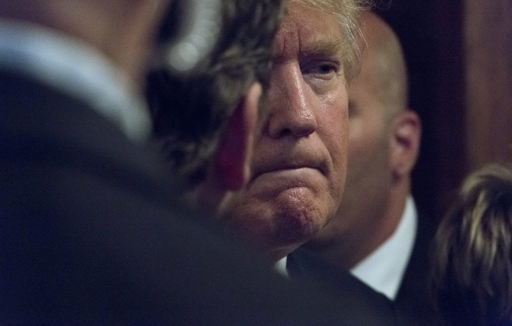 L'image montre un homme avec des cheveux blonds, affichant une expression pensive ou sérieuse. Il est partiellement obscurci par des personnes en arrière-plan, ce qui crée une atmosphère de concentration ou d'intensité. On peut également remarquer des costumes sombres, suggérant un cadre formel.