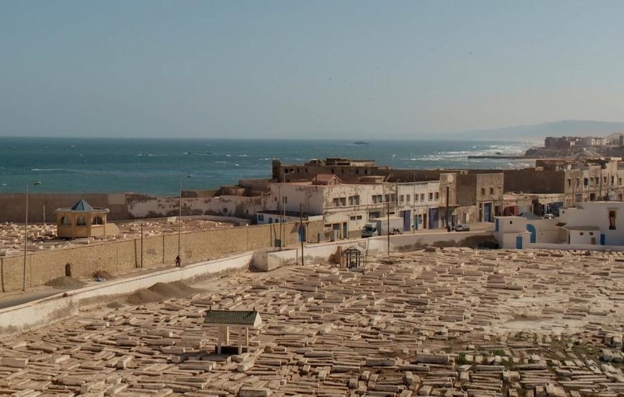 L'image montre un panorama d'une ville côtière, probablement située au bord de la mer Méditerranée. Au premier plan, on peut voir un cimetière avec de nombreuses tombes en pierre, disposées de manière régulière. À l'arrière-plan, il y a des bâtiments blancs, typiques des architectures méditerranéennes, qui bordent la côte. La mer s'étend à l'horizon, avec des vagues visibles, et il semble y avoir une légère brume sur l'eau. La scène dégage une ambiance paisible et sereine, avec une lumière douce évoquant l'heure du soir ou du matin.