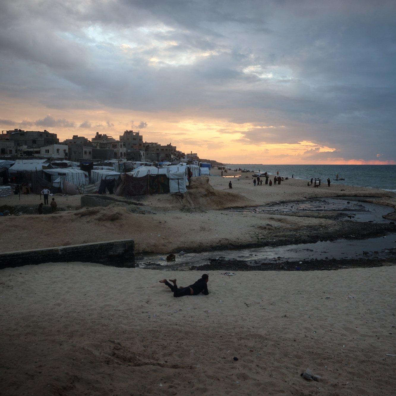 L'immagine mostra una spiaggia al tramonto, con il cielo che presenta nuvole scure e toni arancioni. Sulla spiaggia si trovano diverse tende e strutture, probabilmente utilizzate come rifugi temporanei. L'acqua del mare appare calma, e ci sono alcune persone in lontananza che camminano lungo la riva. In primo piano, si può vedere una parte di spiaggia con sabbia e un piccolo corso d'acqua che scorre. L'atmosfera sembrerebbe evocare un momento di transizione, sia dal punto di vista naturale che umano.