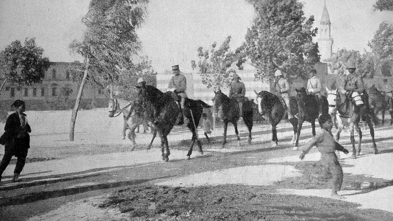 L'image montre un groupe de cavaliers, probablement des soldats, sur des chevaux, défilant sur une route. Il y a des arbres sur les côtés, et au premier plan, on peut voir une personne marchant sur le côté du chemin. À l'arrière-plan, des bâtiments et un clocher sont visibles, suggérant un environnement urbain. L'atmosphère semble historique, peut-être photographiée au début du XXe siècle.