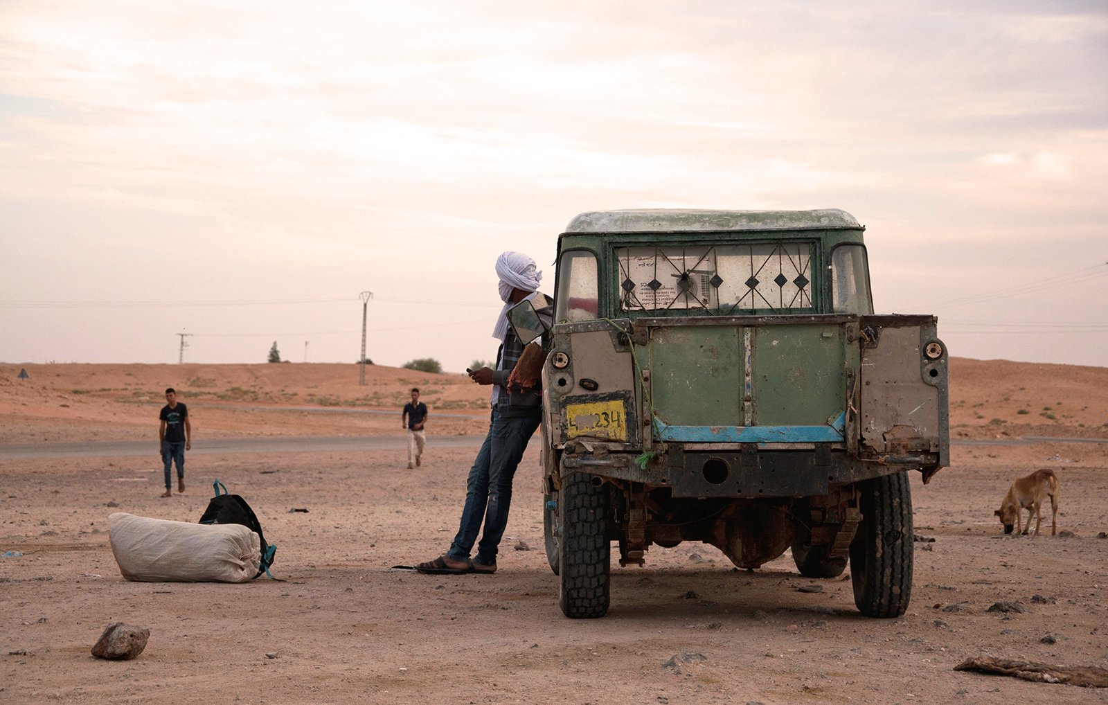 L'image montre une scène en plein air dans un paysage désertique. On peut voir un ancien véhicule utilitaire, avec une carrosserie usée et des fenêtres cassées. Un homme, partiellement visible, se tient à côté du véhicule, portant un turban. En arrière-plan, il y a d'autres personnes, ainsi qu'un chien qui déambule. Le ciel est nuageux, et l'atmosphère semble calme, mais aussi un peu désolée, avec des dunes de sable à l'horizon.