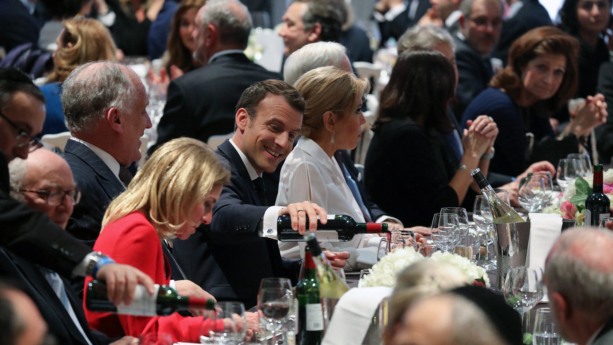 L'image montre une scène de dîner élégant, probablement lors d'un événement officiel. Plusieurs personnes sont assises autour d'une grande table, décorée avec des verres, des bouteilles de vin et des fleurs. Au centre, on aperçoit un homme souriant en train de servir du vin, semblant profiter de l'atmosphère. Les convives sont habillés de manière formelle, et l'ambiance paraît conviviale et animée, avec des conversations visibles parmi les invités.