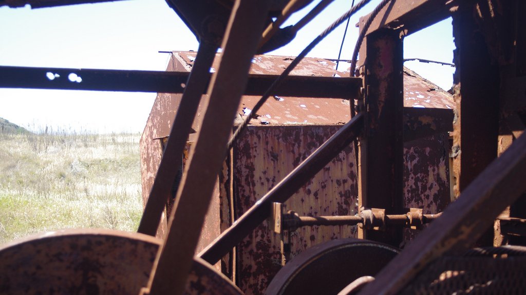 L'image montre une vue d'une structure métallique, probablement une machine ou un équipement abandonné, avec des surfaces rouillées et en décomposition. On peut voir des éléments mécaniques, tels que des poulies ou des engrenages, qui sont partiellement visibles à travers des parties de la machine. En arrière-plan, on distingue un paysage de prairies sèches, ce qui évoque une ambiance d'abandon et de dégradation. La lumière naturelle met en valeur les textures rouillées du métal.