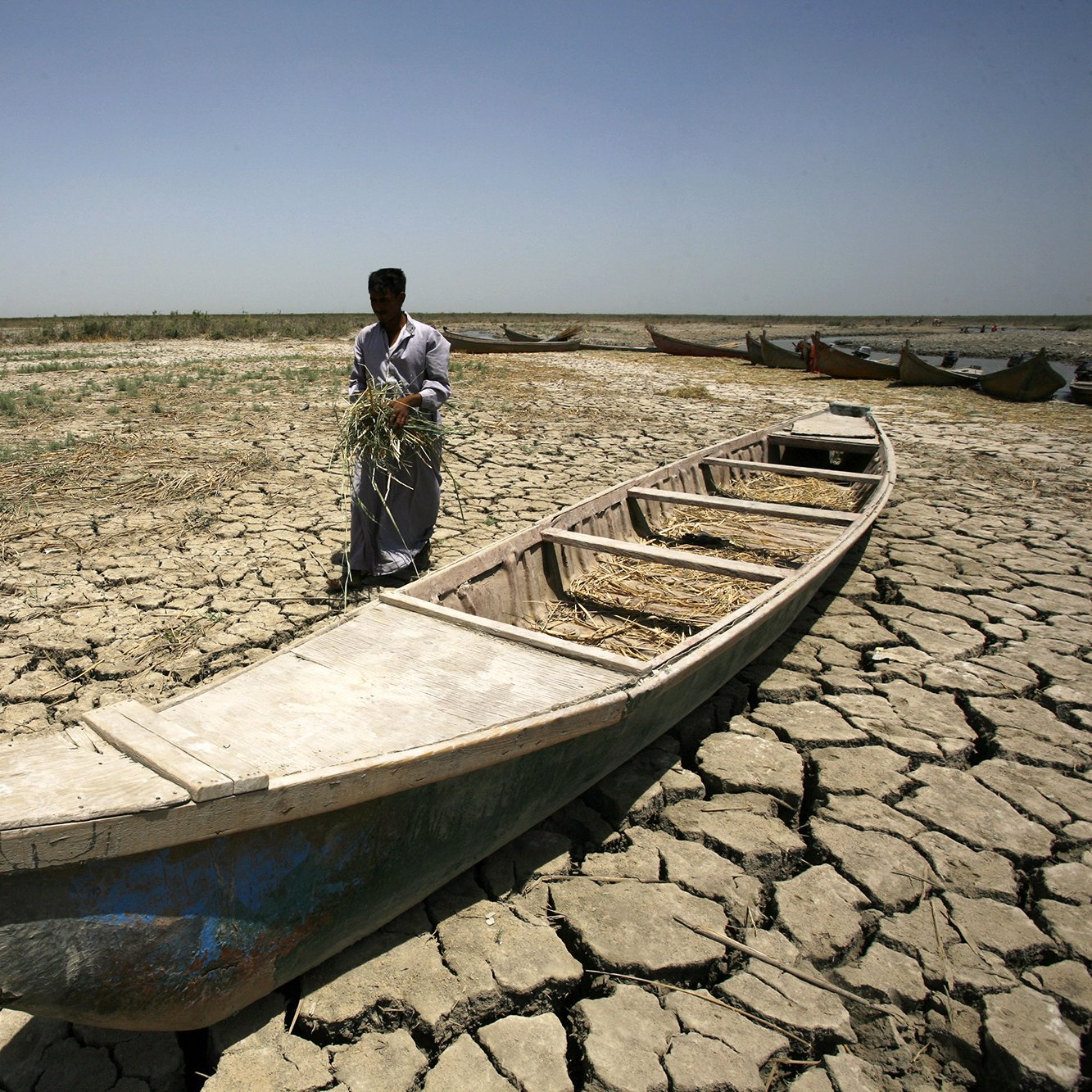 L'image montre un homme debout sur un sol craquelé, typique d'une région aride. Il semble tenir une poignée de végétation, peut-être de l'herbe ou des tiges. À ses pieds se trouve une barque vide, qui est en partie enfouie dans le sol sec. En arrière-plan, on peut apercevoir d'autres barques, mais le paysage aride, sans présence significative d'eau, souligne un environnement désolé et dégradé. Le ciel est dégagé et lumineux, renforçant l'atmosphère de chaleur et de sécheresse.