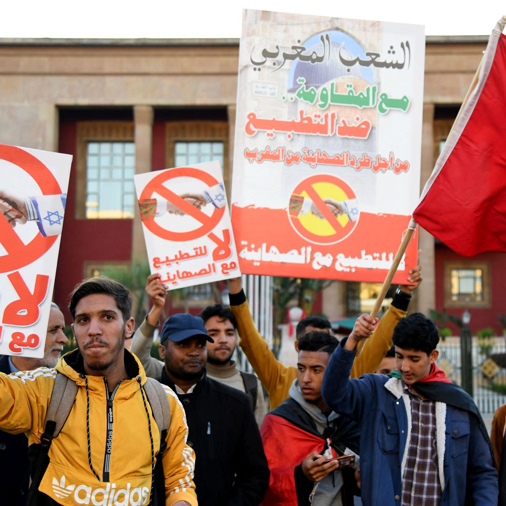 L'image montre un groupe de manifestants tenant des pancartes. Les affiches portent des messages visant à s'opposer à la normalisation des relations avec Israël, avec des symboles qui suggèrent un rejet de cette initiative. Les participants, d'âges divers, expriment leur désaccord à travers des slogans et des gestes. Certains portent des vêtements d'hiver, et l'ambiance semble être celle d'une manifestation pacifique. En arrière-plan, on aperçoit un bâtiment public, probablement lié au gouvernement.