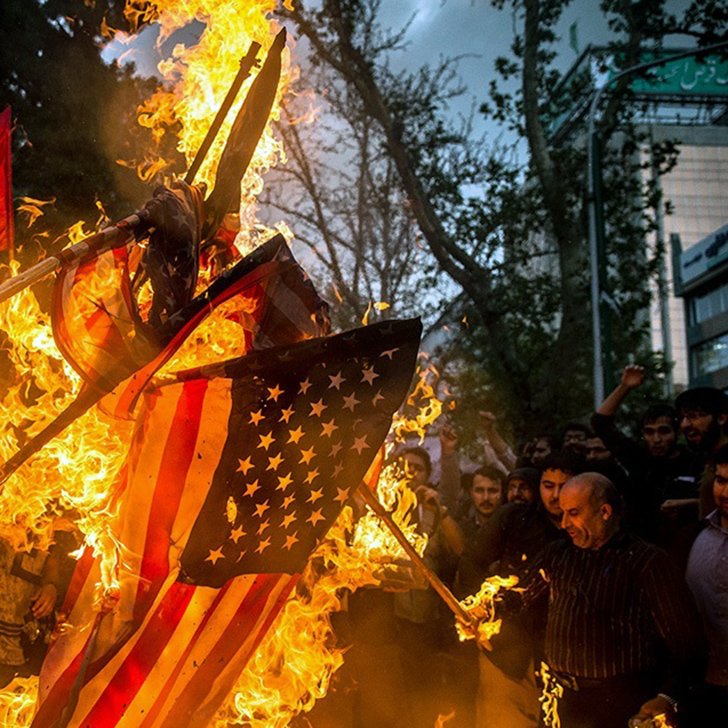 این تصویر نشان‌دهنده یک تجمع اعتراضی است که در آن گروهی از افراد در حال سوزاندن پرچم ایالات متحده هستند. در پس‌زمینه، دیده می‌شود که برخی از افراد دست‌ها را بالا برده و به نشانه اعتراض فریاد می‌زنند. دود و آتش در حال برخاستن از پرچم است و فضا به شدت احساسی و پر از شور و هیجان به نظر می‌رسد. درختان و ساختمان‌هایی نیز در پس‌زمینه وجود دارند که نشان‌دهنده مکان شهری این رویداد است.