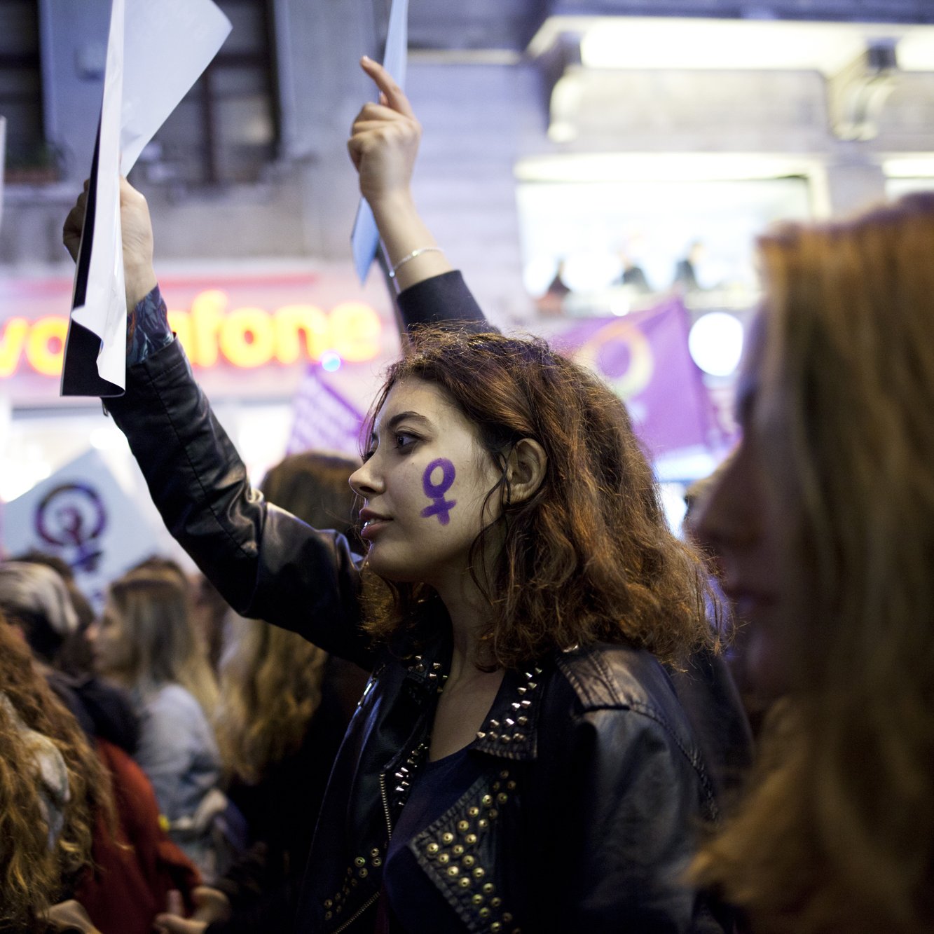 L'image montre un groupe de personnes participant à une manifestation. Au premier plan, une jeune femme avec des cheveux bouclés et un maquillage marqué, porte une veste en cuir et tient un morceau de papier ou une affiche. Son visage est décoré d'un symbole féminin en violet, qui se retrouve également sur d'autres bannières en arrière-plan. L'atmosphère semble être celle d'un rassemblement pour les droits des femmes, soulignant la solidarité et l'engagement des participants.