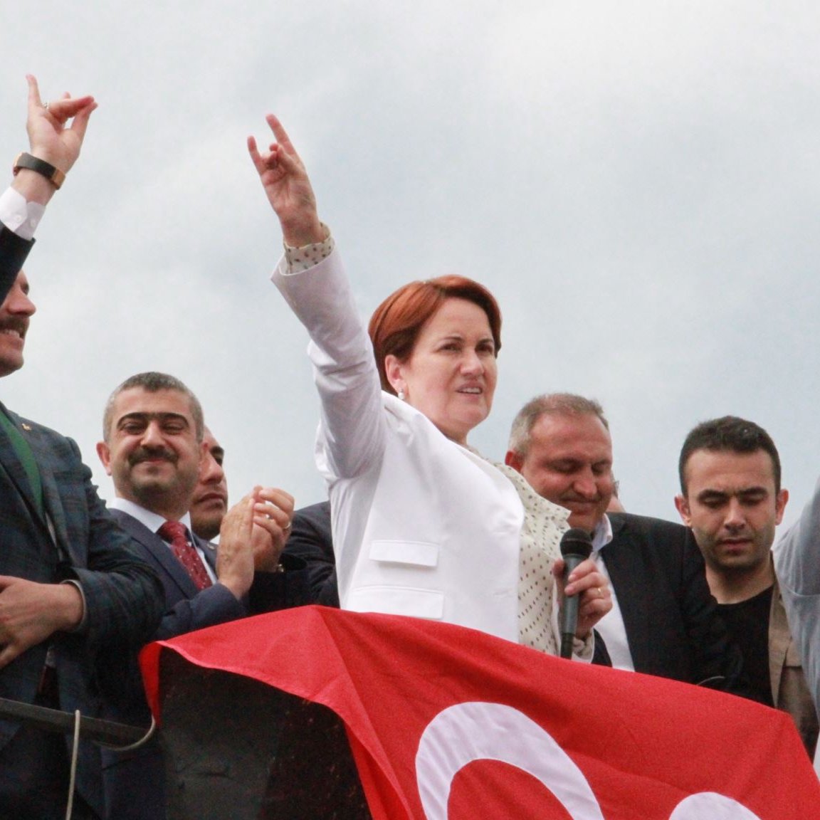 L'image montre un groupe de personnes sur une scène, avec une femme au centre qui lève le bras en signe de victoire tout en tenant un micro. Elle porte une tenue blanche et semble s'adresser à la foule. Derrière elle, plusieurs hommes, dont certains applaudissent, sont visibles. Une grande bannière rouge est accrochée sur la scène, ajoutant une touche de couleur à l'image, avec un ciel nuageux en arrière-plan. L'ambiance semble festive et militante.