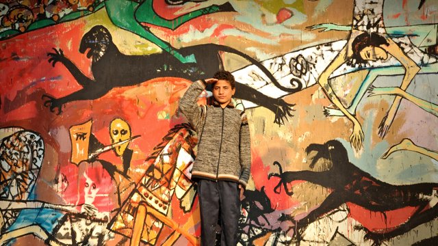 L'image montre un jeune garçon en train de faire un salut militaire devant un mur coloré, sur lequel sont peintes diverses figures stylisées et abstraites. Le mur présente des scènes dynamiques, avec des motifs de monstres et d'êtres humains, créant un contraste frappant avec le garçon qui porte un manteau gris et un pantalon sombre. Son expression et sa posture semblent témoigner de confiance ou de détermination. Les couleurs vives et les formes exagérées de la fresque attirent l'œil et ajoutent une dimension artistique à la scène.
