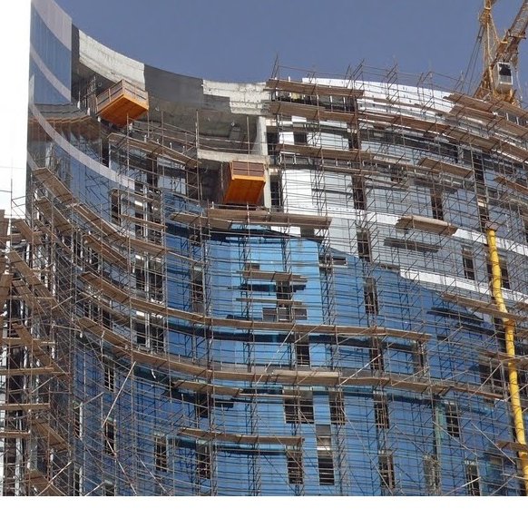 L'image montre un bâtiment en construction. On peut voir une structure moderne avec des façades en verre, entourée d'un échafaudage en métal. Plusieurs niveaux sont visibles, et des éléments de construction, comme des grues, sont présents pour faciliter les travaux. Le ciel est dégagé, ce qui indique une belle journée.