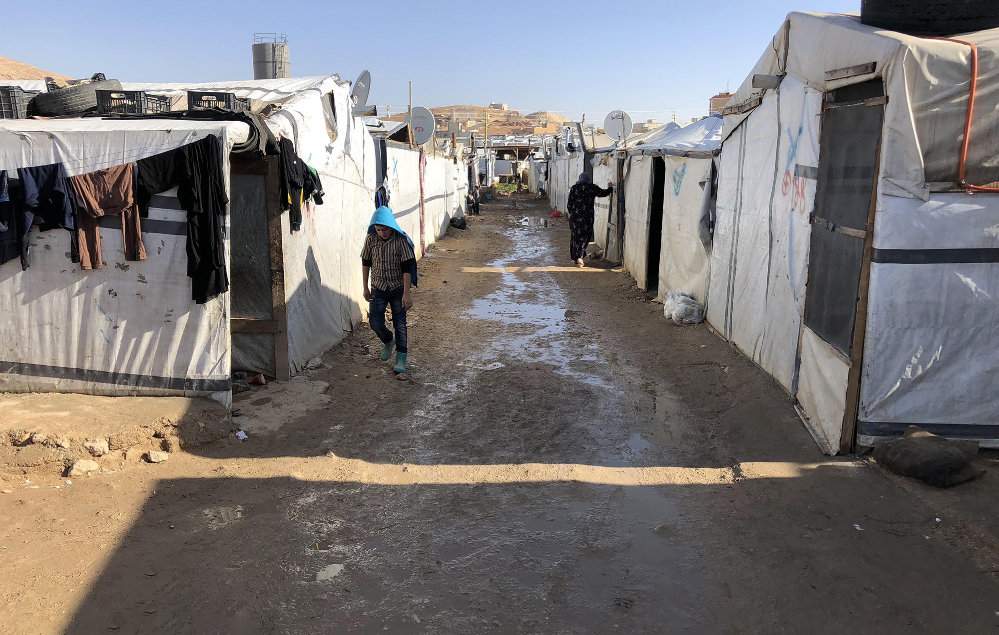 L'image montre un camp de réfugiés, avec des tentes blanches et des structures temporaires alignées des deux côtés d'un chemin étroit. Le sol semble boueux, probablement à cause de la pluie. On peut voir des vêtements accrochés à l'extérieur des tentes. Une personne, vêtue de manière simple, marche le long du chemin central. L'environnement semble rudimentaire et surpeuplé, avec des signes de vie quotidienne.