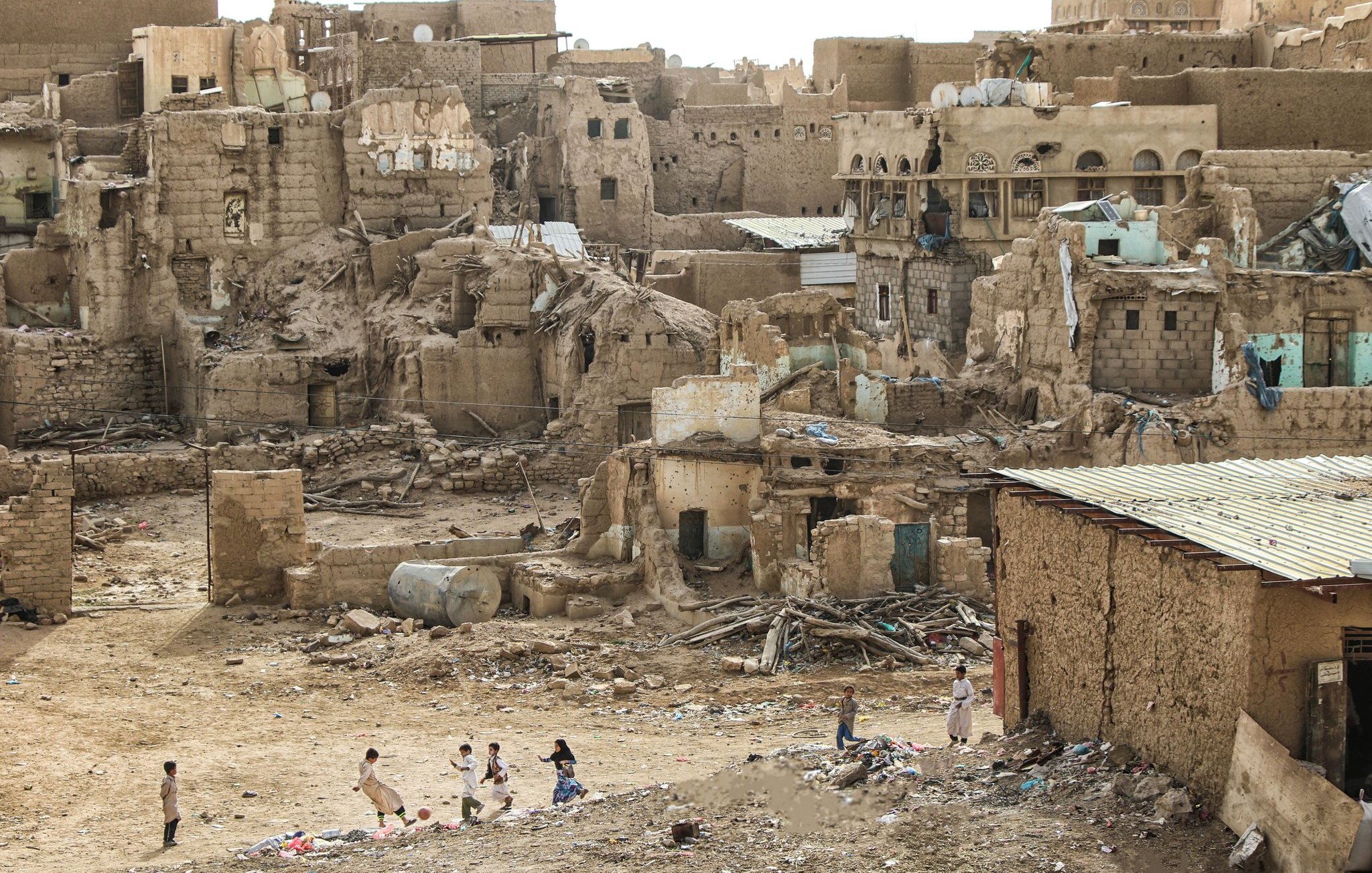 تصویر یک منطقه مخروبه و قدیمی را نشان می‌دهد که ساختمان‌ها و دیوارهای فرسوده و آجرهای شکسته در آن دیده می‌شود. در foreground، چند کودک در حال بازی هستند. زمین خشک و بی‌حاصل به نظر می‌رسد و بقایای ساخت و ساز و زباله‌ها در اطراف پراکنده شده‌اند. این منظر حس نابودی و تخریب را منتقل می‌کند و نمایی از زندگی در یک منطقه آسیب‌دیده را به نمایش می‌گذارد.