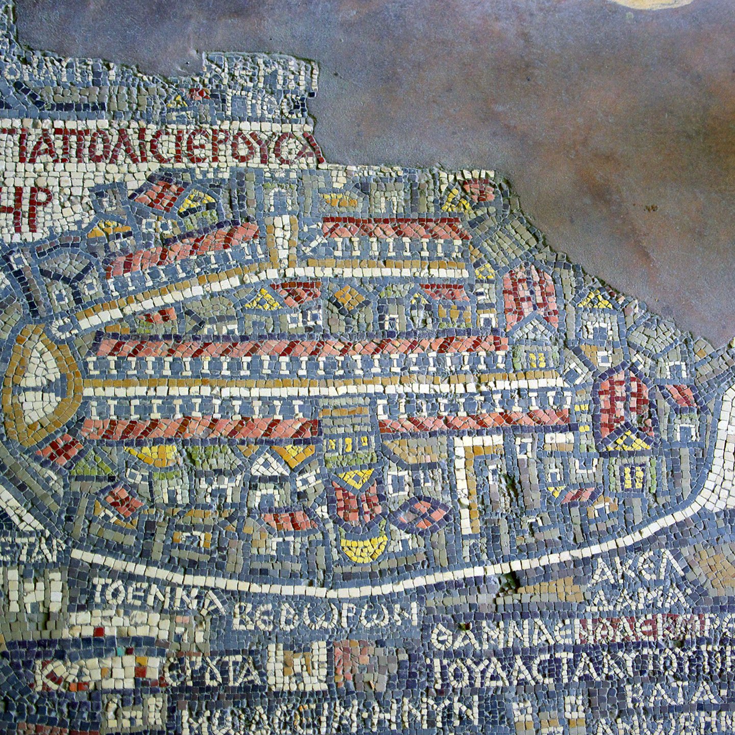 Cette image montre un ancien mappemonde représentant la ville de Jérusalem, réalisée en mosaïque. On y distingue des motifs géométriques et des bâtiments stylisés, disposés de manière à évoquer la topographie de la ville. Les inscriptions en grec ancien ajoutent des détails sur les lieux et les quartiers. La mosaïque utilise une palette de couleurs variées, créant un effet visuel riche et complexe, typique de l'art byzantin.