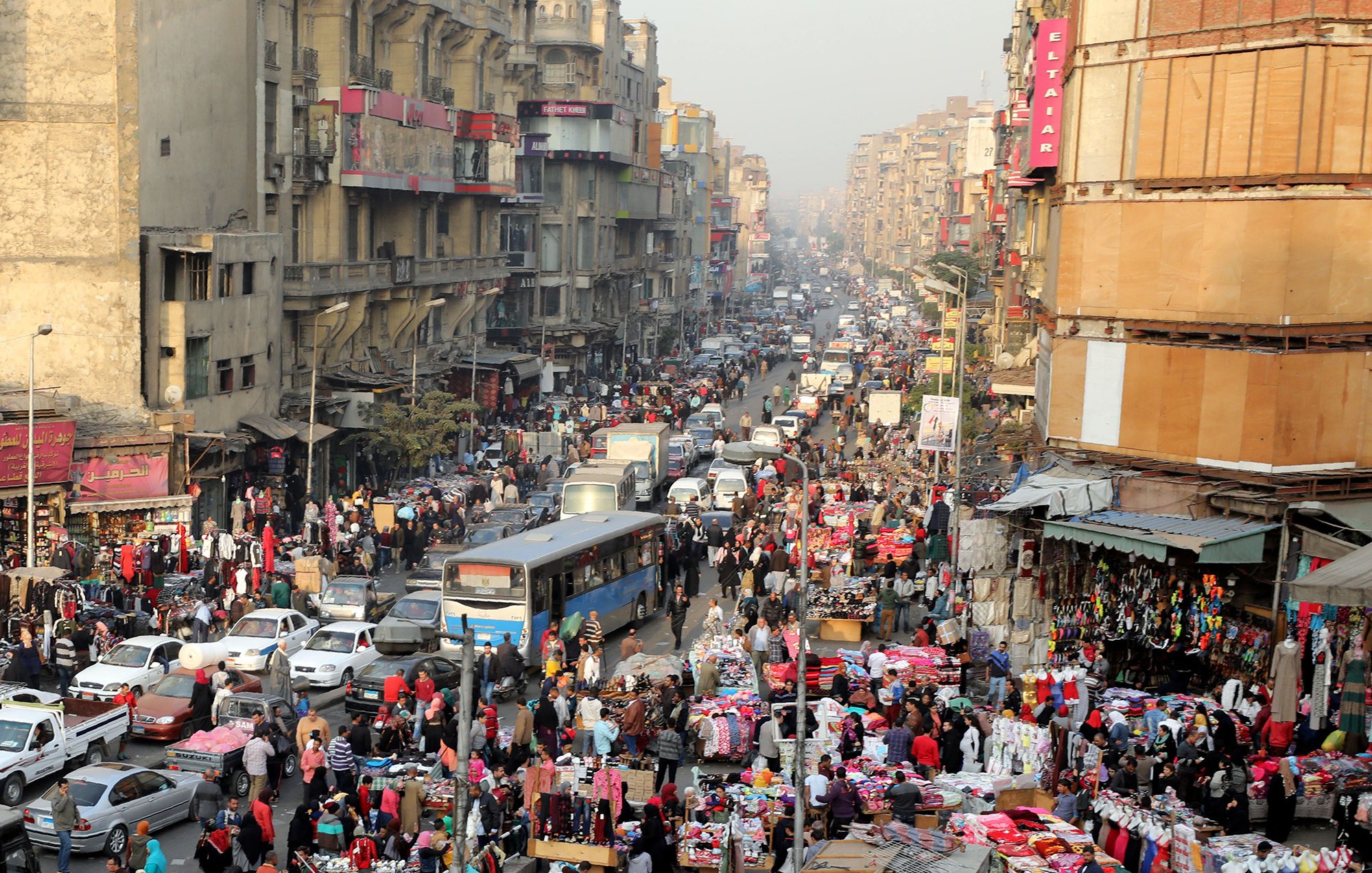 L'image montre une scène animée d'un marché urbain bondé, probablement dans une grande ville. On peut voir des foules de personnes se mêlant aux voitures dans une rue étroite. Les étals colorés sont remplis de divers produits, tels que des vêtements et des accessoires. Les bâtiments environnants semblent anciens et plusieurs niveaux sont visibles, avec des enseignes visibles. L'atmosphère paraît vivante et dynamique, typique des marchés populaires dans les grandes agglomérations.