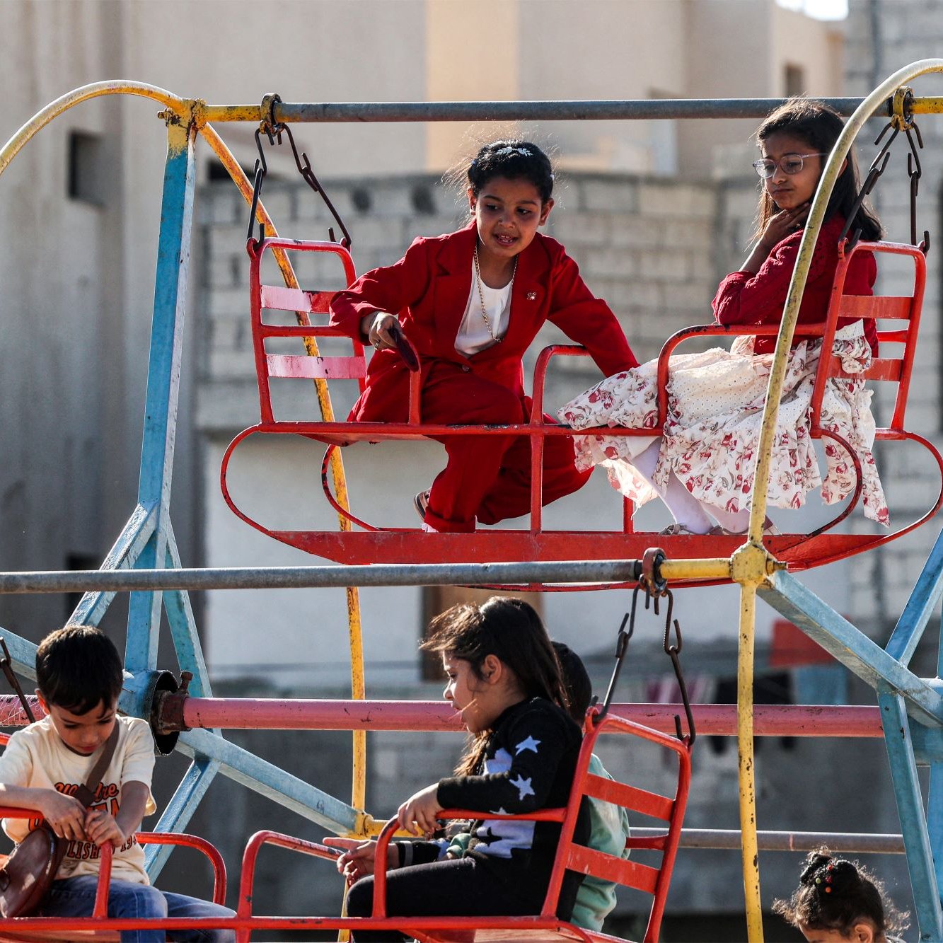 L'image montre des enfants jouant sur une grande structure de jeux colorée. On voit des balançoires fixées à une plateforme, dont certains enfants sont assis. Les enfants affichent des expressions joyeuses, profitant de leur temps de jeu. La scène se déroule dans un espace ouvert, avec des bâtiments en arrière-plan. Les vêtements des enfants sont variés, ajoutant de la vivacité à l'ensemble. L'atmosphère semble joyeuse et familiale.