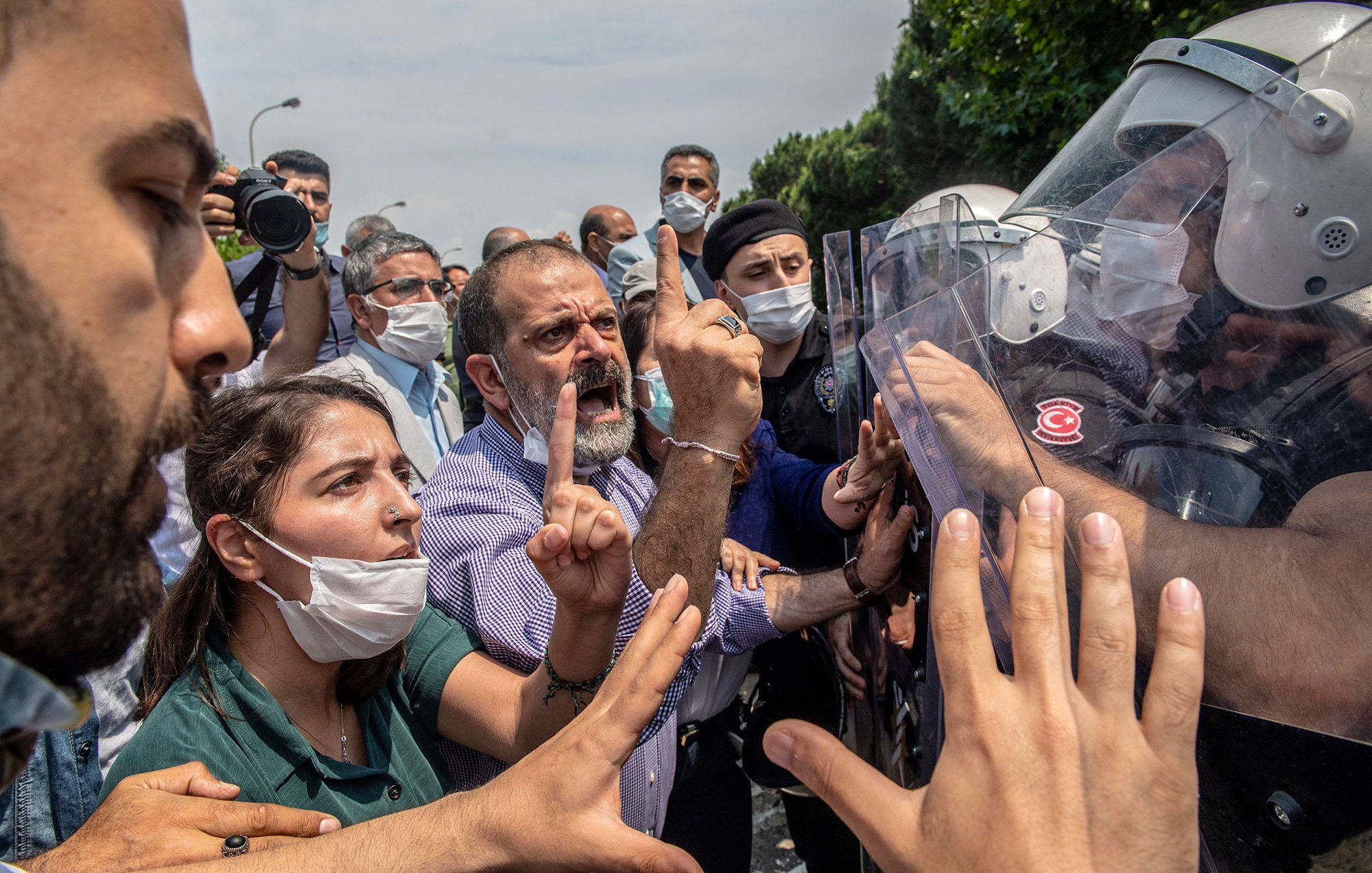 L'image montre une scène de tension entre des manifestants et des forces de l'ordre. Au premier plan, plusieurs personnes expriment leur désaccord, avec des gestes vifs et des expressions de colère ou de frustration. Certaines personnes portent des masques, probablement en raison de préoccupations sanitaires. En arrière-plan, des membres des forces de l'ordre, équipés de casques et de boucliers, semblent faire face aux manifestants. L'environnement est urbain, et le ciel est partiellement nuageux. L'atmosphère est chargée d'émotion et de conflit.