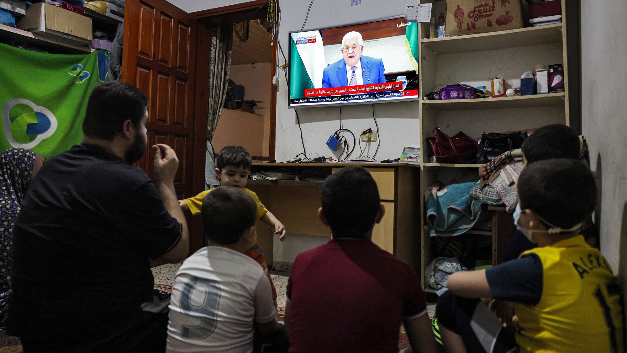 L'immagine mostra un gruppo di bambini seduti a terra, rivolti verso un televisore che trasmette un programma. Un adulto, probabilmente un genitore o un parente, è accanto a loro. La stanza sembra essere piccola e modesta, con mobili semplici e un'atmosfera informale. Sullo schermo si vede un uomo che parla, probabilmente un leader o un personaggio pubblico. I bambini appaiono curiosi e coinvolti nella visione.