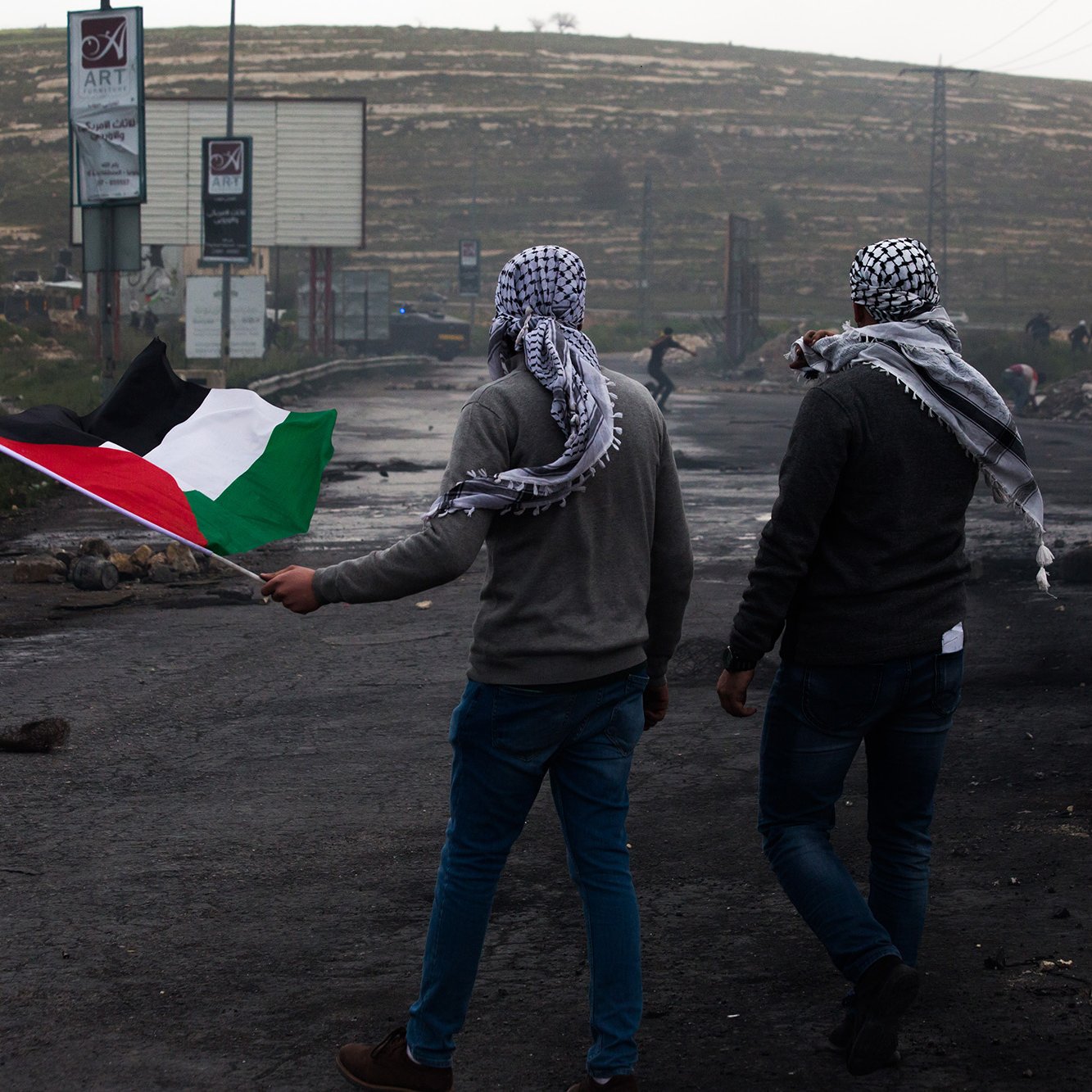 L'image montre deux hommes marchant sur une route, chacun portant un keffieh. L'un d'eux tient un drapeau palestinien, qui est composé de bandes noire, blanche, verte et rouge. L'arrière-plan présente des panneaux publicitaires, des débris et une ambiance brumeuse. Les hommes semblent se diriger vers une direction, symbolisant probablement une manifestation ou une action de revendication.