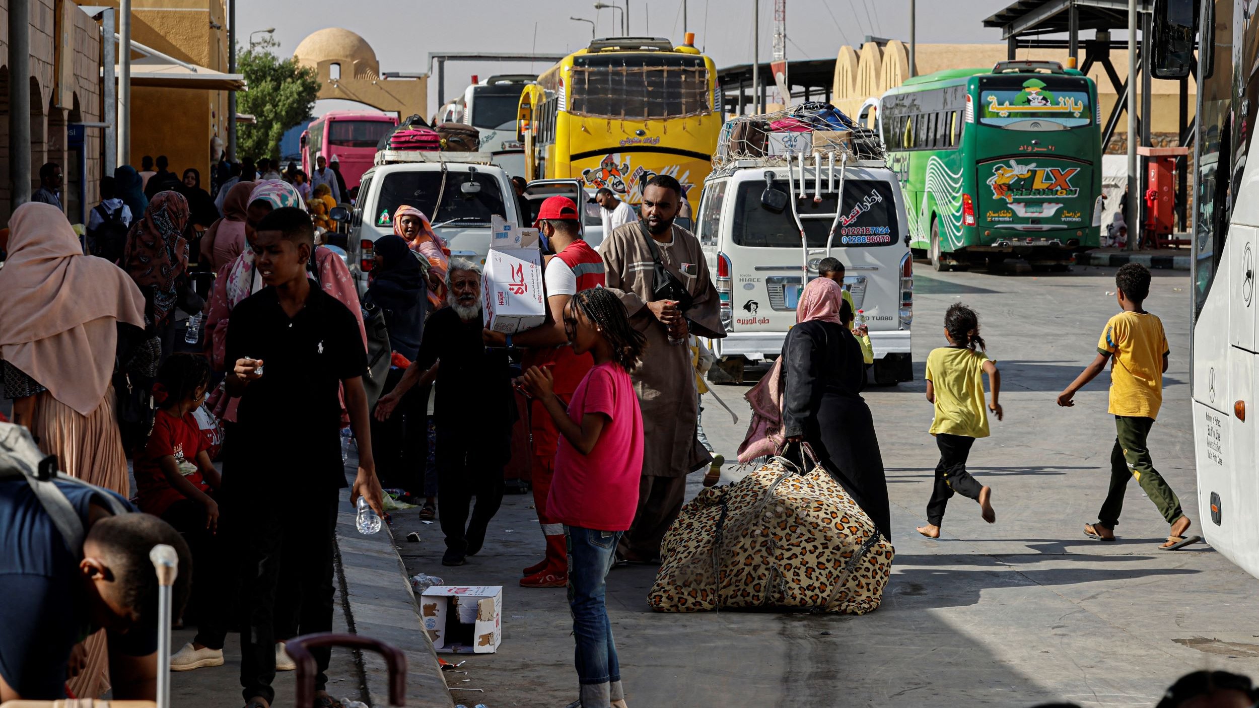 La imagen muestra una estación de autobuses donde hay un gran movimiento de personas. Se puede ver a varias familias y grupos de personas caminando entre los autobuses, algunos con maletas y bultos. Hay un ambiente de actividad, con adultos y niños, algunos consumiendo bebidas y otros llevando objetos. En el fondo, se aprecian varios autobuses de diferentes colores, indicando que podría ser un lugar de transporte importante. La escena refleja un momento de desplazamiento y espera.