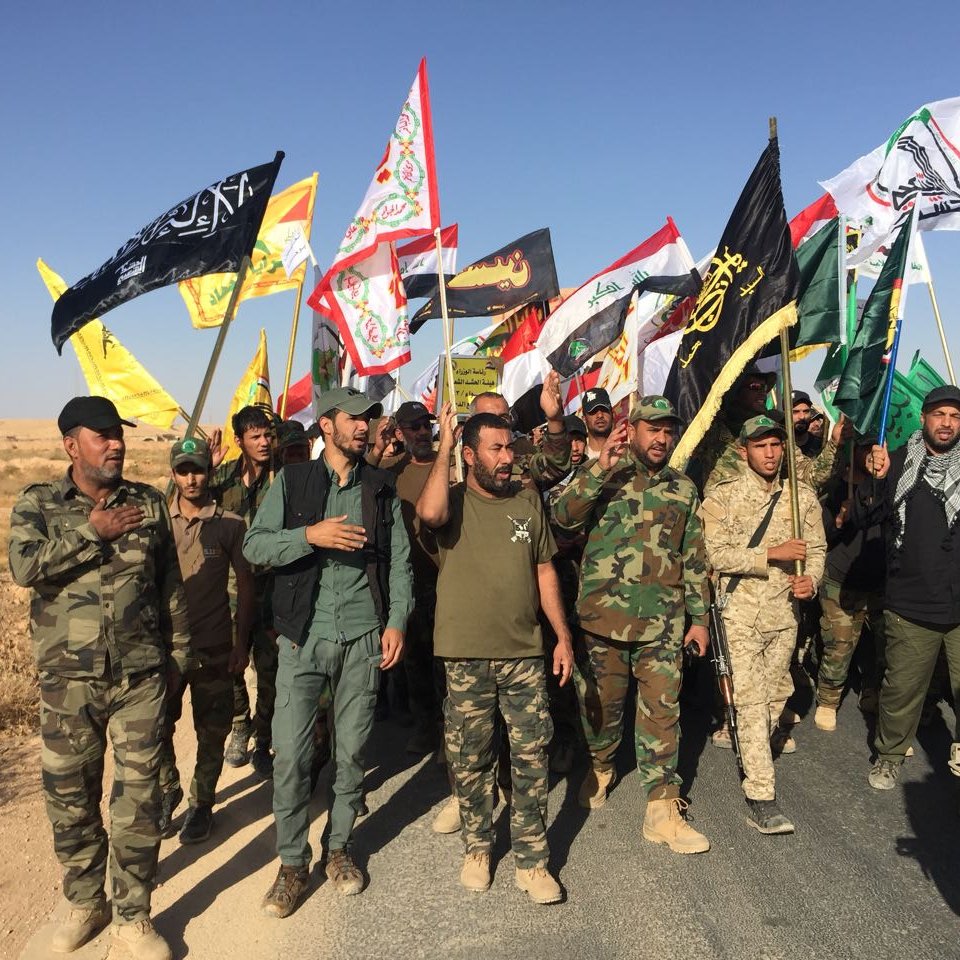 L'image montre un groupe de personnes participant à une manifestation ou une procession. Elles portent des uniformes militaires et tiennent des drapeaux de différentes couleurs et motifs, représentant probablement des groupes ou des mouvements politiques ou religieux. Le paysage environnant est désertique, avec un ciel bleu clair. Les participants semblent engagés et déterminés, marchant ensemble sur une route.