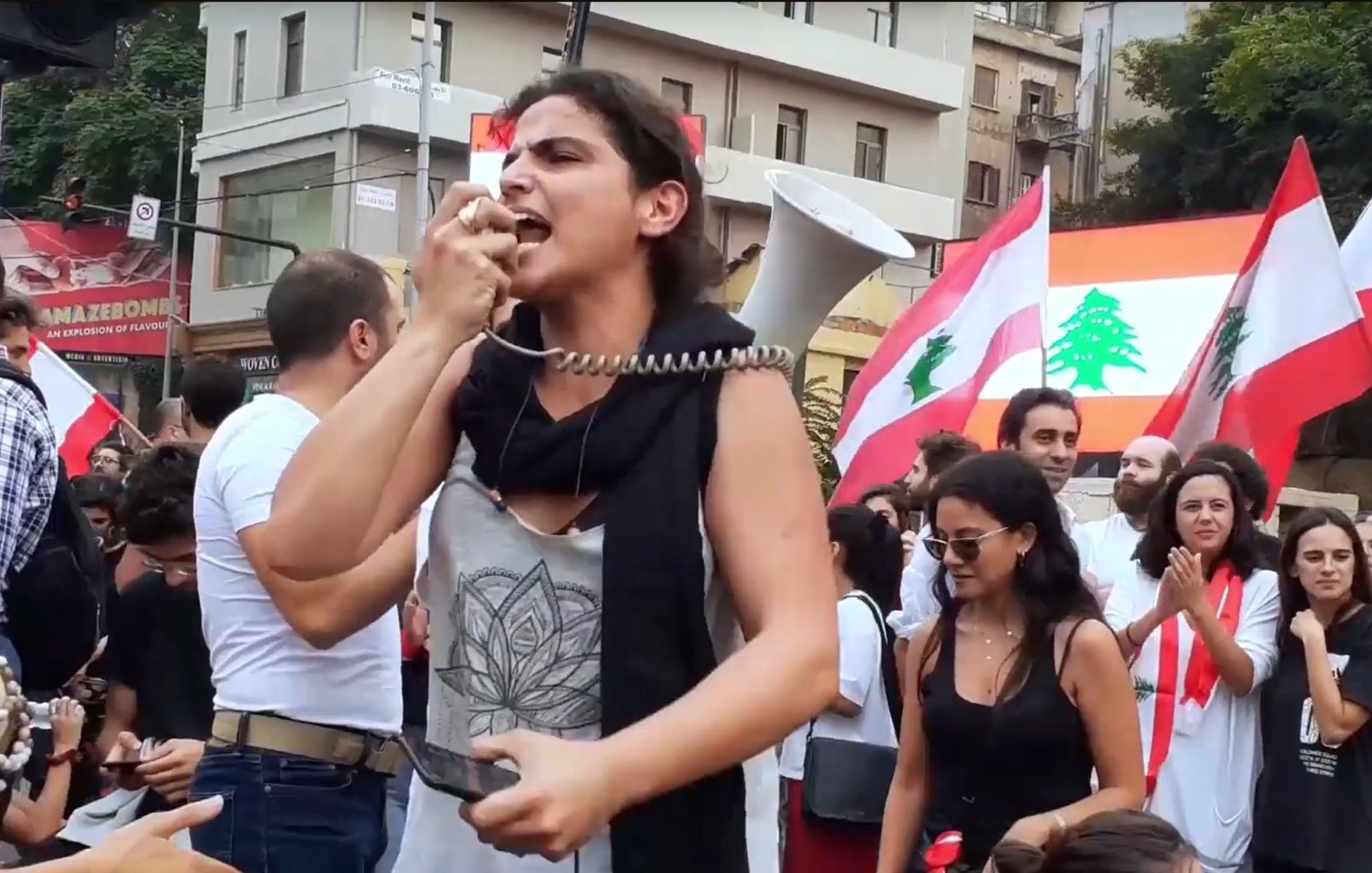 Foule en manifestation, femme au mégaphone, drapeaux libanais en arrière-plan.
