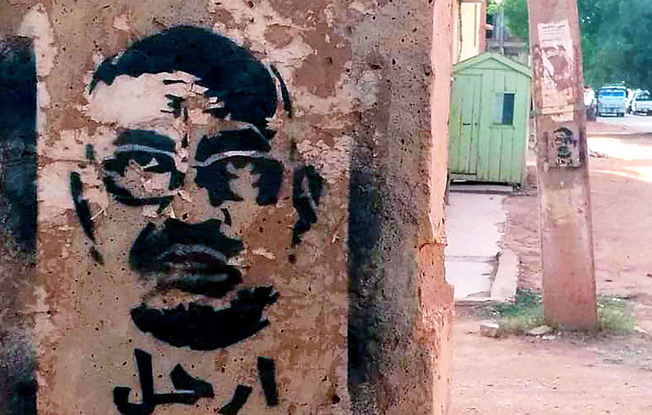 L'image montre un graffiti sur un mur, représentant un visage stylisé d'un homme avec des lunettes. Le dessin est en noir, sur un fond de béton clair. En arabe, le mot "ارحل" est inscrit, ce qui signifie "pars" ou "sors". L'environnement semble urbain, avec des bâtiments et une route visible en arrière-plan.