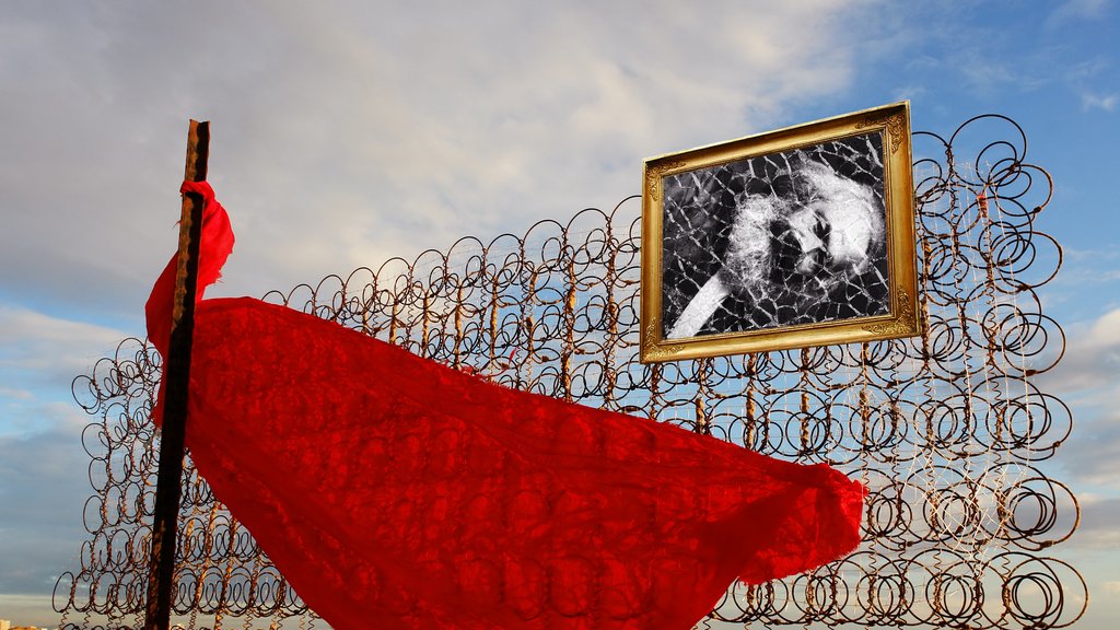 L'immagine mostra una scena poetica e surreale. Al centro, un drappo rosso fluttua nel vento, appendendosi a una struttura metallica composta da spirali di ferro. Dietro il drappo, vi è una cornice dorata che contiene una fotografia in bianco e nero, probabilmente di un soggetto significativo. Il cielo sullo sfondo è ampio, con nuvole sparse che creano un'atmosfera suggestiva. L'insieme evoca temi di libertà, memoria e contrasto tra materiali ruvidi e il delicato tessuto rosso.