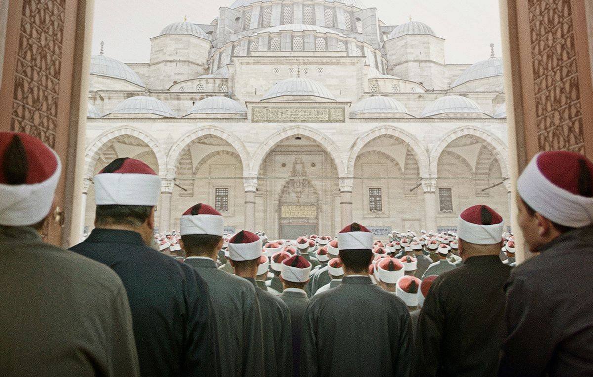 L'image montre un grand groupe de personnes, probablement des religieux ou des fidèles, qui se tiennent debout, tournés vers un imposant bâtiment architectural qui semble être une mosquée. Les hommes portent des vêtements similaires, souvent dans des tons sombres, et certains portent des turbans ou des chapeaux à pompons rouges. L'ambiance semble solennelle et respectueuse, suggérant un moment de prière ou de rassemblement communautaire. Les détails architecturaux de la mosquée, avec ses coupoles et arches majestueuses, ajoutent à l'imposante scène.