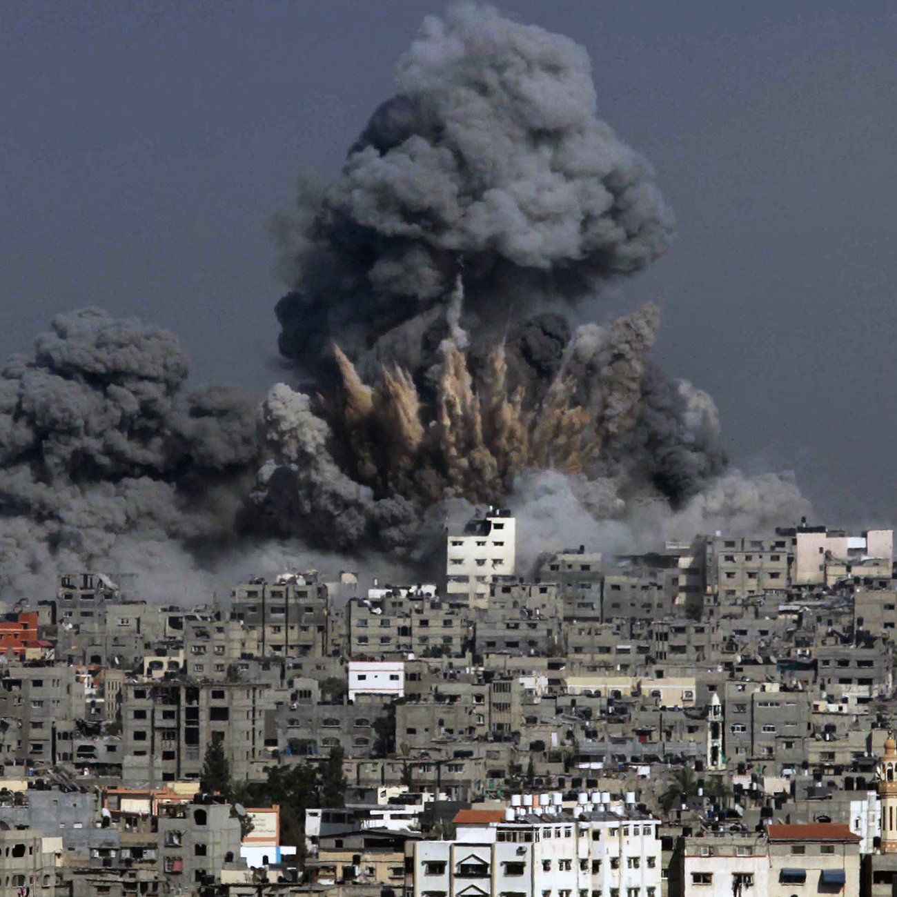 L'image montre une scène de destruction où des colonnes de fumée noire s'élèvent dans le ciel, indiquant qu'une explosion a eu lieu. Les bâtiments sont présents au premier plan, témoignant d'une zone urbaine, et semblent avoir subi des dommages. Le contraste entre la couleur grise de la fumée et les structures environnantes évoque une atmosphère de chaos et de conflit.