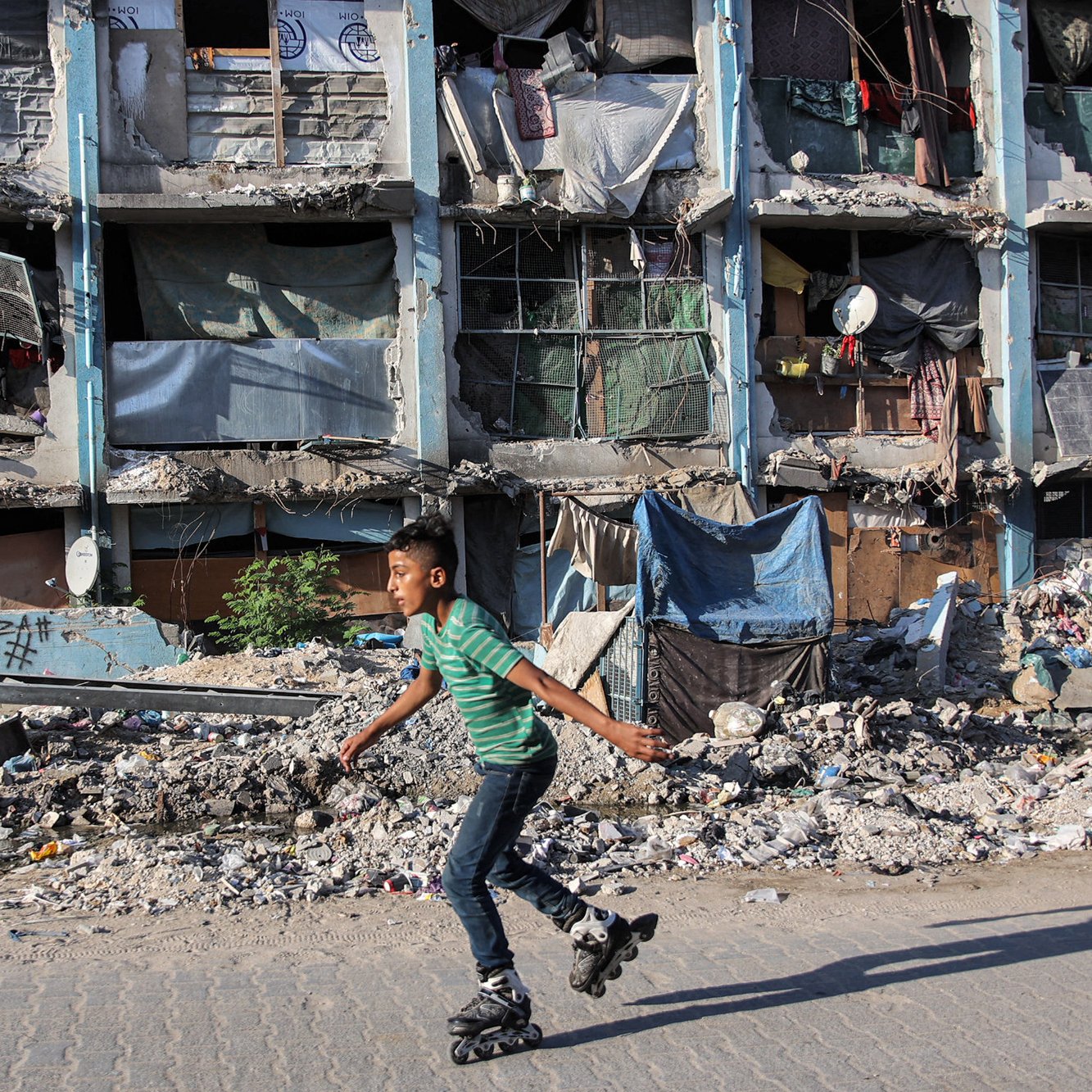 L'immagine ritrae un ragazzo che pattina su una superficie dura, indossando un paio di pattini a rotelle. Sullo sfondo si possono vedere edifici danneggiati e fatiscenti, con finestre rotte e tende appese. L'area circostante è caratterizzata da detriti e spazzatura, che suggeriscono una situazione di degrado urbano. Nonostante l'ambiente difficile, il ragazzo sembra impegnato e concentrato mentre pattina.
