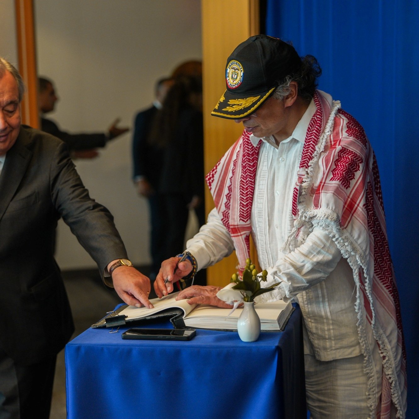 Two men are at a table; one is signing a book while the other observes. A blue backdrop is behind them.