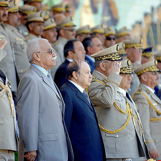 L'image montre un groupe de personnalités militaires en uniforme, saluant avec respect. Ils se tiennent debout en rangs, avec un décor cérémoniel en arrière-plan. Les hommes portent des uniformes ornés de médailles et de chapeaux militaires, et l'atmosphère semble officielle et solennelle, probablement lors d'une célébration ou d'un événement commémoratif. Des fleurs sont visibles au premier plan, ajoutant à l'ambiance formelle de la scène.