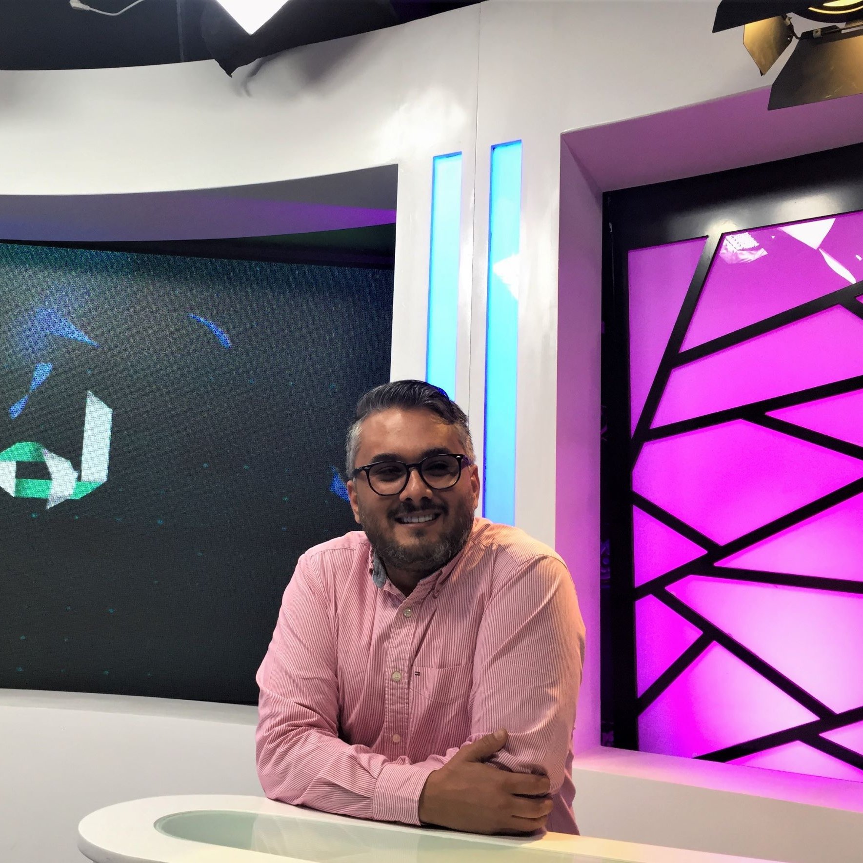 L'immagine mostra un uomo sorridente seduto davanti a una scrivania in uno studio televisivo. Indossa una camicia rosa e presenta un aspetto rilassato e accogliente. Dietro di lui si possono notare schermi che mostrano un logo o un testo in arabo, mentre l'illuminazione dello studio è accentuata da luci colorate, in particolare tonalità di viola e blu. L'ambiente sembra moderno e professionale, tipico di una trasmissione televisiva.