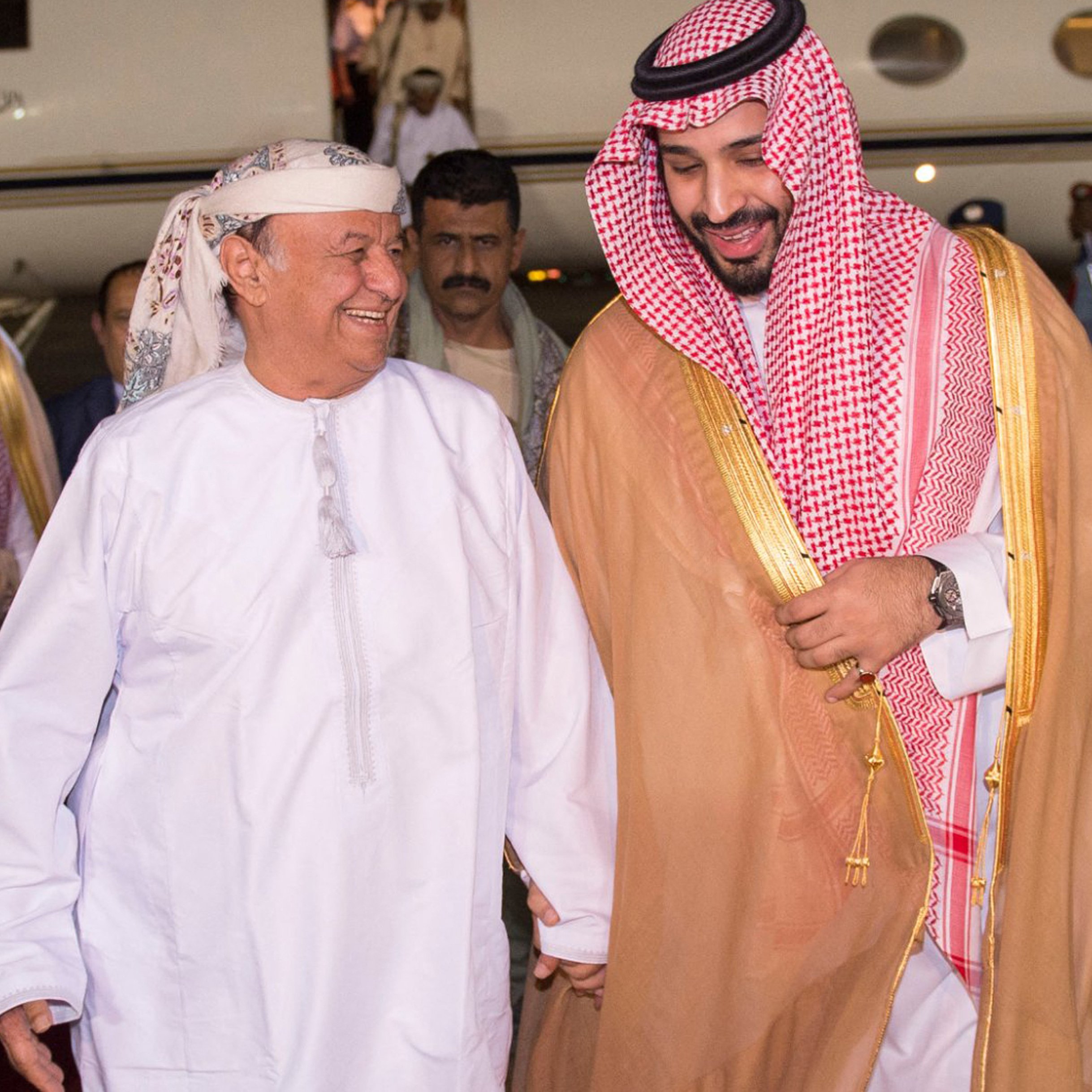 في الصورة، يمكن رؤية رجلين يتحدثان معاً بمرح. يرتدي أحدهما زياء تقليدية عربية مكونة من جبة بيضاء وعقال، بينما يرتدي الآخر الملابس السعودية التقليدية، تشمل العباءة والكوفية. يظهر خلفهم طائرة خاصة، مما يشير إلى وصولهم إلى مكان محدد. تبدو الأجواء مفعمة بالترحاب والود.