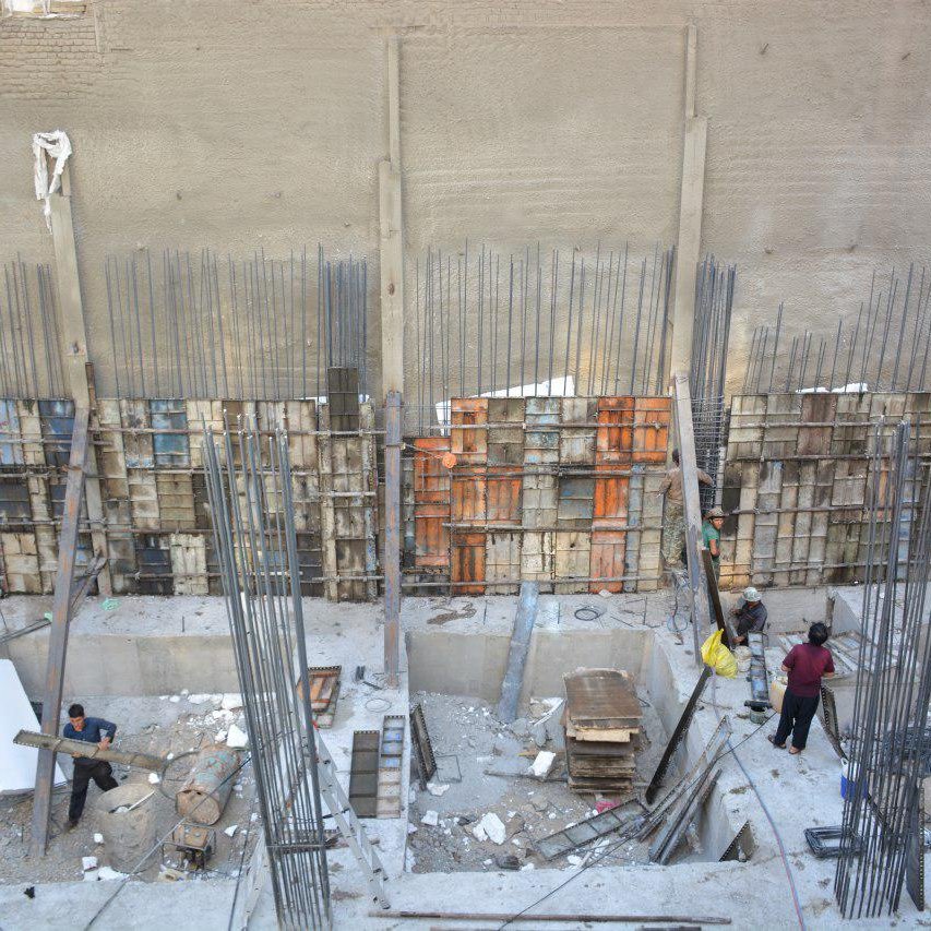 L'image montre un chantier de construction. On peut y voir des ouvriers qui travaillent dans une excavation, entourés de barres d'acier et de matériaux de construction. Les murs en béton sont partiellement visibles, avec des panneaux de coffrage en bois et en métal disposés verticalement. L'environnement semble être en cours de développement, avec une variété d'outils et de matériaux éparpillés autour de l'espace de travail.