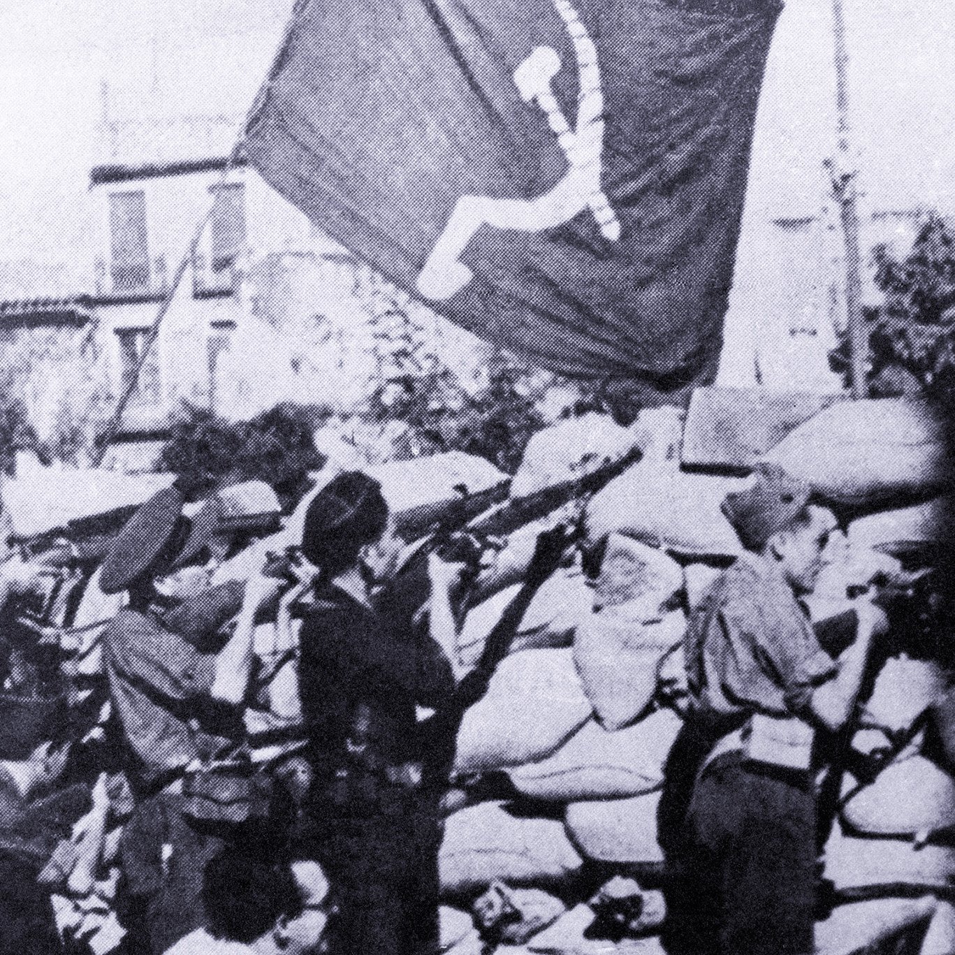 Cette image en noir et blanc montre un groupe de soldats ou de miliciens en position de tir, probablement lors d'un conflit. Ils se trouvent derrière un mur de sacs de sable, tout en tenant des fusils. En arrière-plan, des drapeaux avec un symbole communiste flottent, ce qui suggère un contexte politique et historique marqué par des idéologies de gauche. L'ambiance de la scène semble intense et sérieuse, indiquant un moment de bataille ou de lutte.