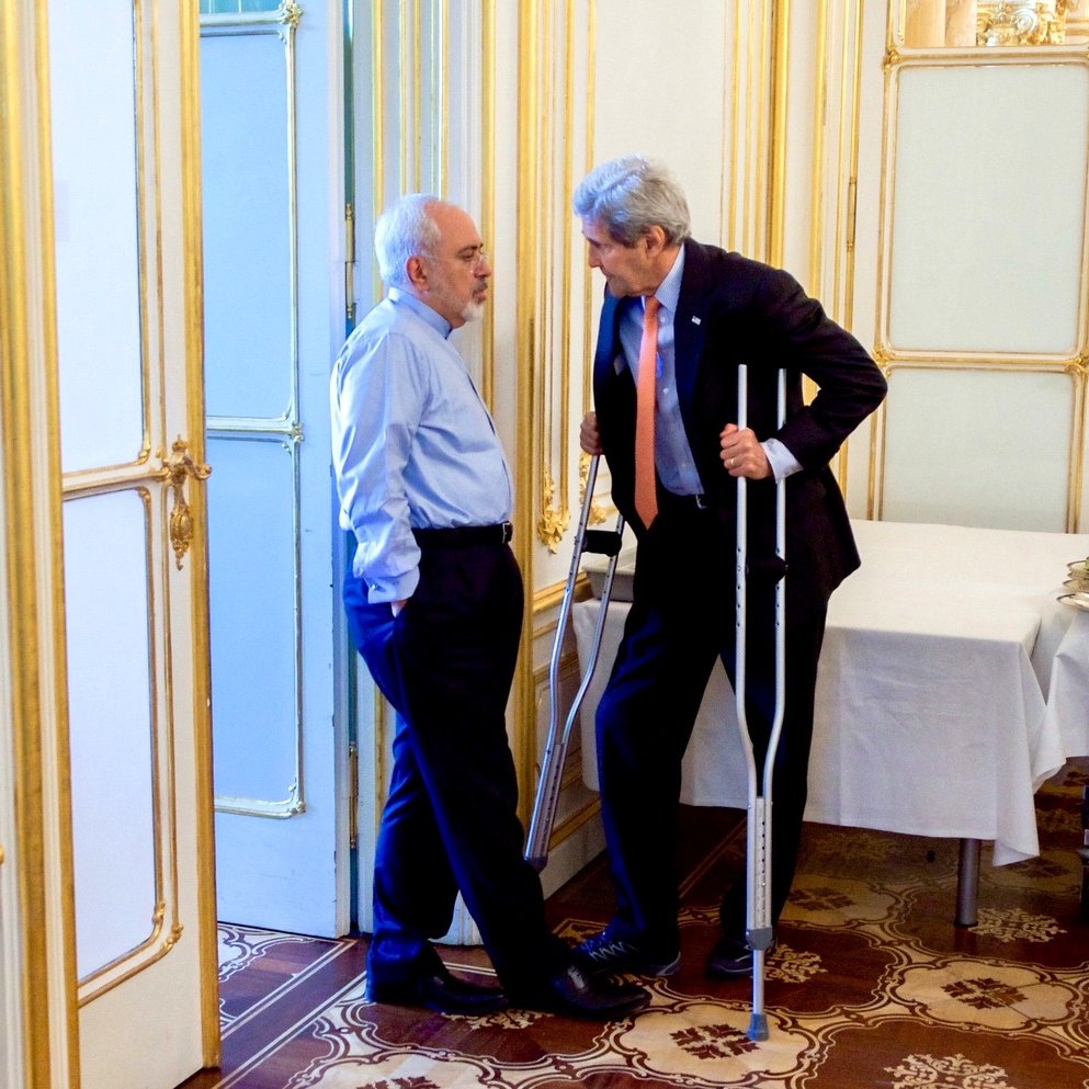 L'image montre deux hommes en train de discuter dans un environnement intérieur. L'un des hommes porte une chemise bleue et semble écouter attentivement, tandis que l'autre, utilisant des béquilles, parle avec animation. L'arrière-plan est décoré avec des moulures dorées et il y a une table couverte d'une nappe blanche. L'atmosphère semble informative et sérieuse.