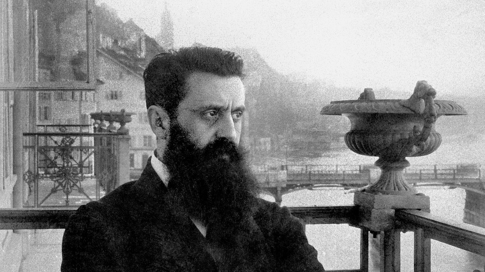L'image montre un homme avec une barbe fournie, portant un costume sombre. Il est debout sur un balcon orné de balustrades métalliques, avec un regard pensif et sérieux. En arrière-plan, on aperçoit des montagnes et un plan d'eau, suggérant un paysage pittoresque. L'atmosphère est nostalgique, renforcée par le ton en noir et blanc de la photographie, ajoutant une dimension historique à la scène.