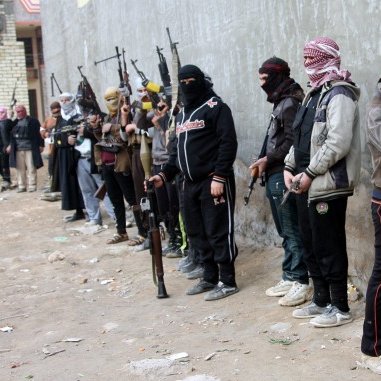 L'image montre un groupe de personnes armées, se tenant alignées le long d'un mur. Elles portent des vêtements sombres et des couvertures sur la tête, ce qui masque leurs visages. Chacune d'elles tient une arme à feu, et certaines adoptent des postures prêtes au combat. L'environnement semble urbain, avec des bâtiments en arrière-plan et un terrain sale. L'atmosphère générale est tendue et évoque un contexte de conflit ou de violence.