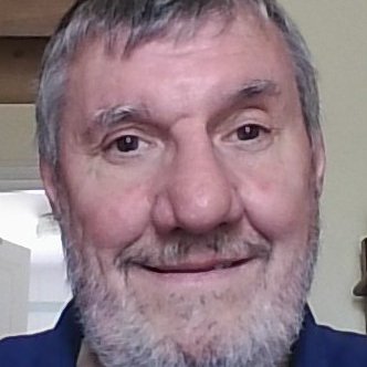 Homme souriant, cheveux gris, barbe fournie, portant un polo bleu. Arrière-plan flou.