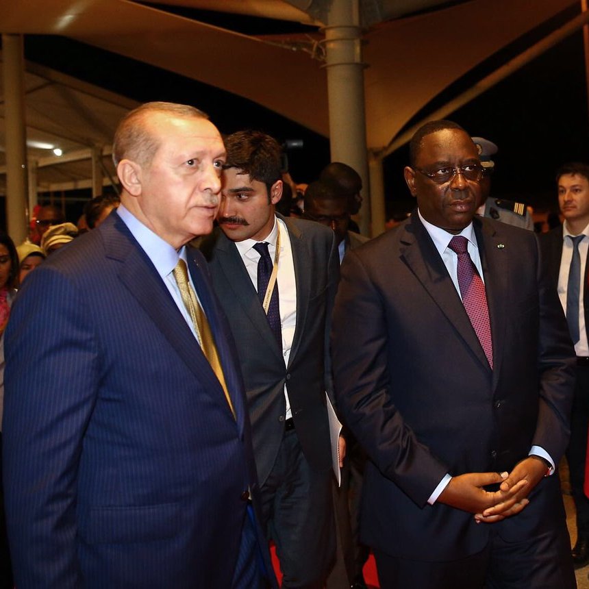 L'image montre deux hommes en costume, marchant ensemble dans un cadre officiel. L'un d'eux semble être un dirigeant turc, tandis que l'autre représente le Sénégal. Ils sont entourés de plusieurs personnes en arrière-plan, et un garde d'honneur en uniforme rouge se tient à proximité. L'atmosphère paraît sérieuse et formelle, typique d'une rencontre diplomatique.