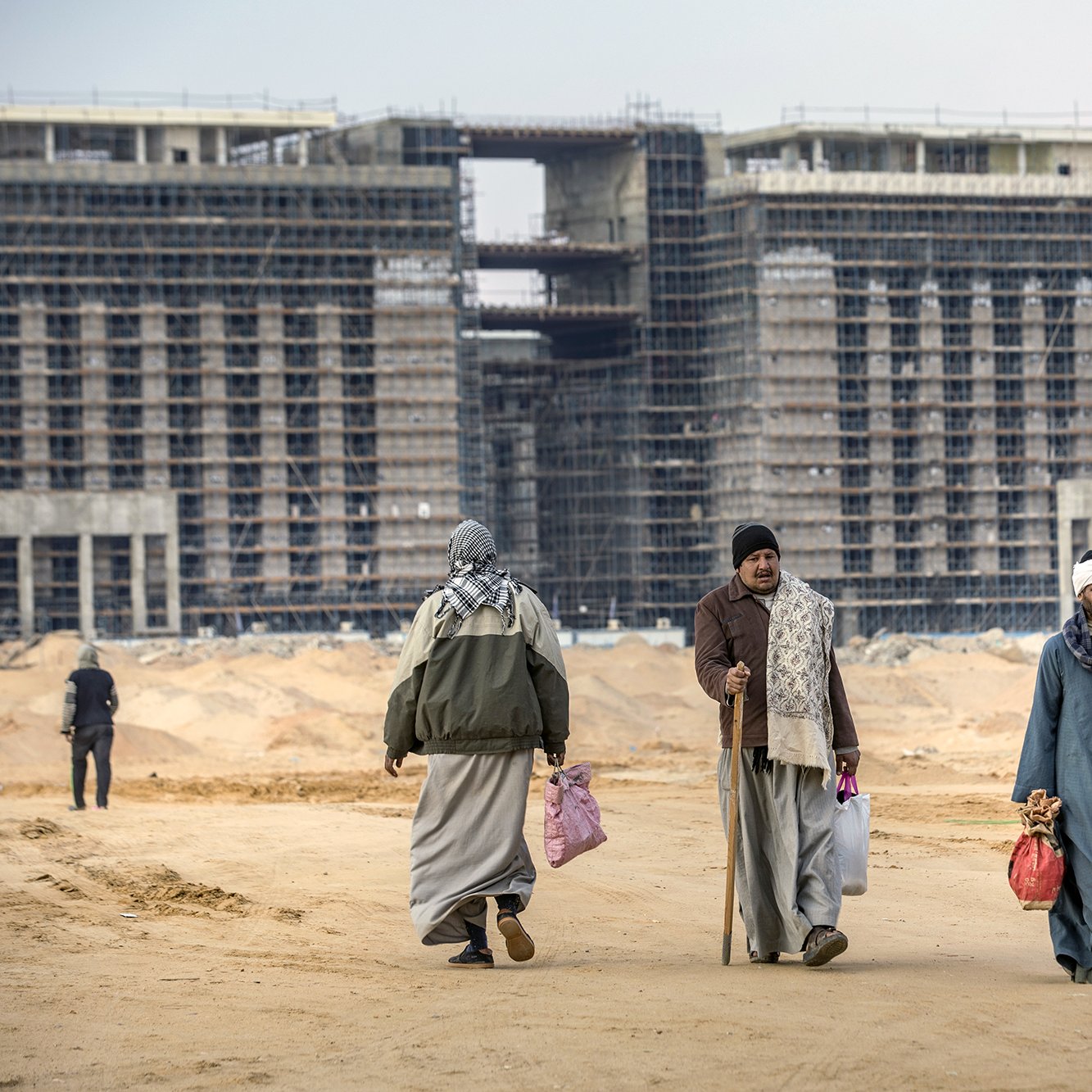 در تصویر، گروهی از مردان در محیطی بیابانی و ساختمانی در حال حرکت هستند. پشت سر آن‌ها، ساختمان‌های در حال ساخت دیده می‌شود که نمایی خالی و ناتمام دارند. افراد با لباس‌های سنتی، برخی با چوبدست و کیسه‌هایی در دست، در حال رفت و آمد در جاده‌ای شن‌زاری هستند. آسمان کمی ابری به نظر می‌رسد و فضایی نسبتاً خنک و خاموش برقرار است.