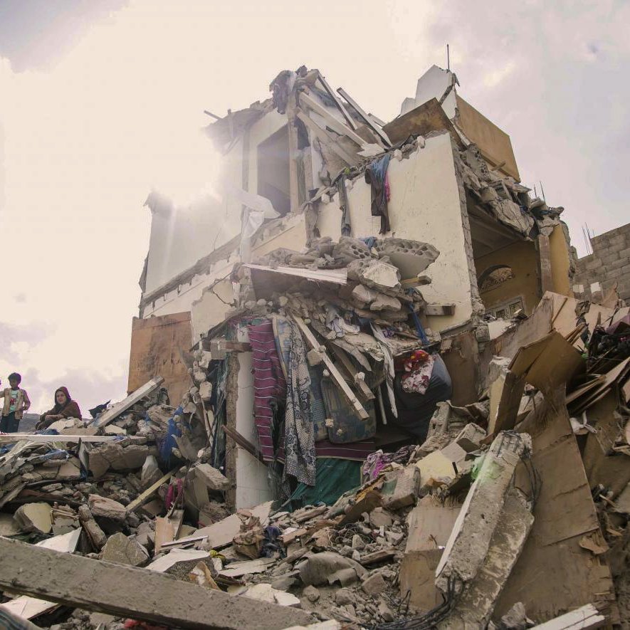 L'image montre un bâtiment effondré, témoignant des dégâts causés par un tremblement de terre ou une catastrophe similaire. Les débris sont éparpillés autour, et on peut voir des personnes s'approchant des ruines. La lumière du soleil perçoit à travers les restes du bâtiment, ajoutant une atmosphère dramatique à la scène de destruction. Les couleurs sont ternes et le paysage donne une impression de désolation et de tristesse face à la situation.