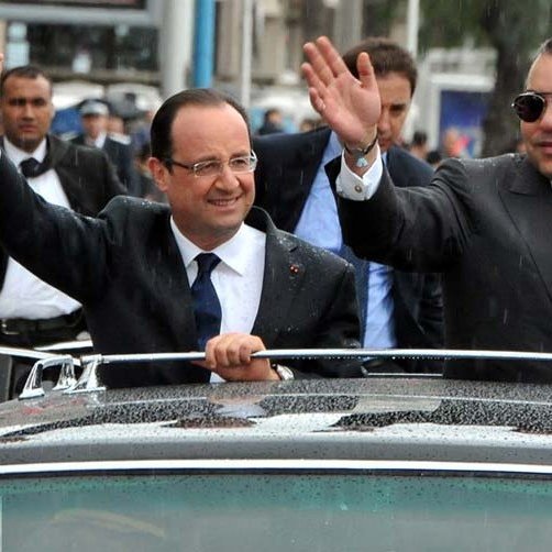 L'image montre deux hommes qui semblent être dans une voiture décapotable. L'un d'eux, à gauche, porte un costume sombre avec une cravate, tandis que l'autre à droite porte un costume avec des accessoires distinctifs. Ils saluent la foule avec la main, semblant sourire et être en bonne humeur. En arrière-plan, il y a des personnes et des voitures, probablement lors d'un événement officiel ou d'une visite. Le temps semble être pluvieux, avec des gouttes d'eau visibles sur la voiture.