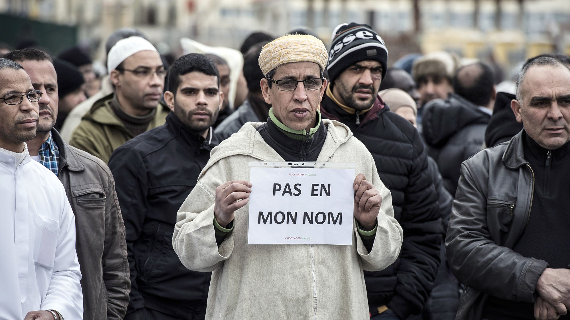 L'image montre un groupe de personnes rassemblées, dont certaines portent des vêtements d'hiver. Au premier plan, un homme se distingue en tenant une pancarte sur laquelle est écrit « PAS EN MON NOM ». Son expression est sérieuse et déterminée. Les autres personnes autour de lui semblent également concentrées sur la situation. L'ambiance générale de la scène suggère une manifestation ou un rassemblement pour une cause particulière.