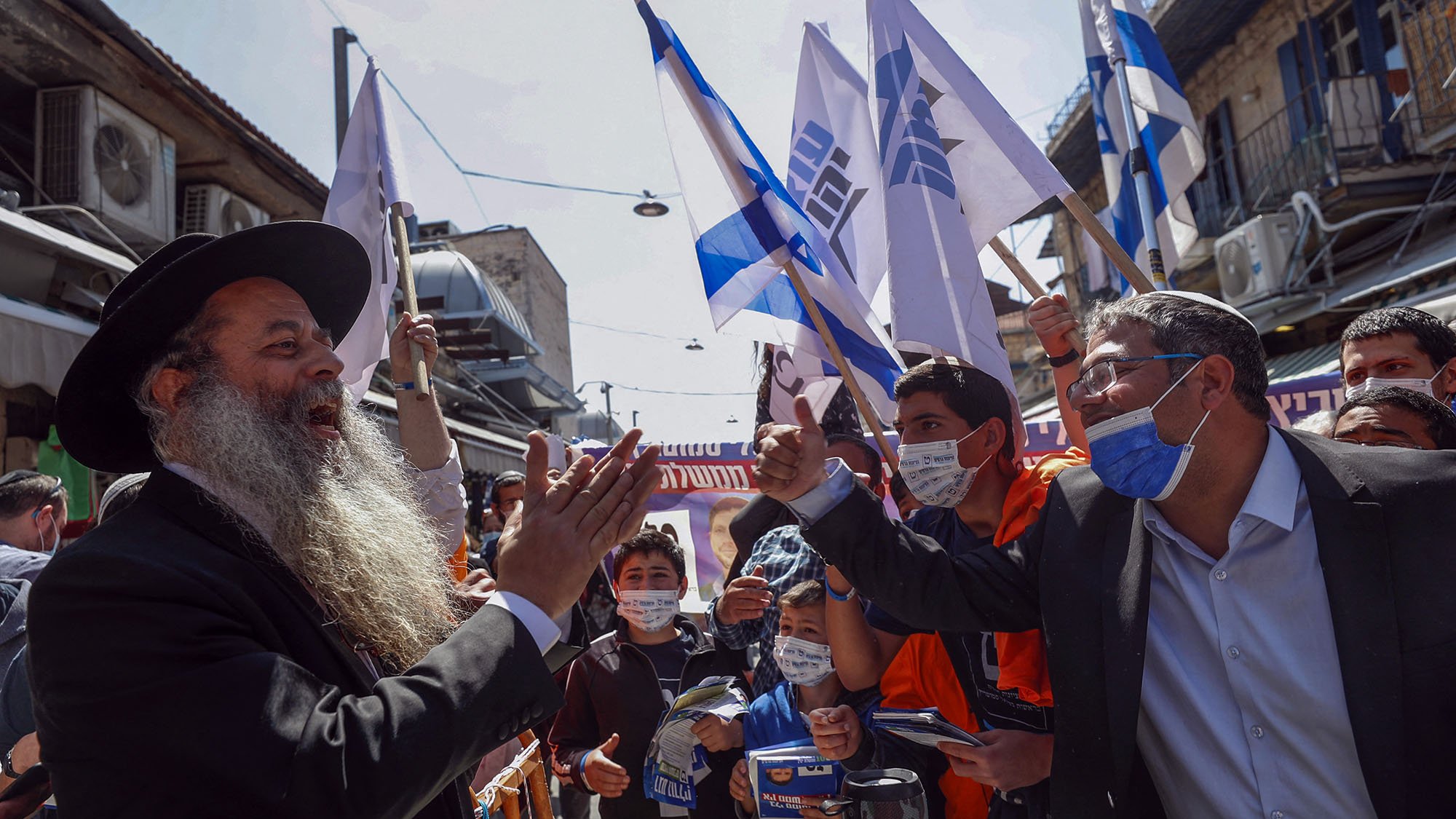 در این تصویر، مردی با ریش بلند و کلاه سیاه در حال سخنرانی و برقراری تعامل با جمعیتی است که پرچم‌های اسرائیل را در دست دارند. افراد حاضر در تصویر، که تعدادی از آن‌ها ماسک به صورت دارند، به نظر می‌رسد در حال ابراز حمایت یا اعتراض هستند. فضا به شدت شلوغ و پرانرژی به نظر می‌رسد و به وضوح نشان‌دهنده یک رویداد اجتماعی یا سیاسی است. ساختارها و تجسس‌های شهری نیز در پس‌زمینه دیده می‌شود.